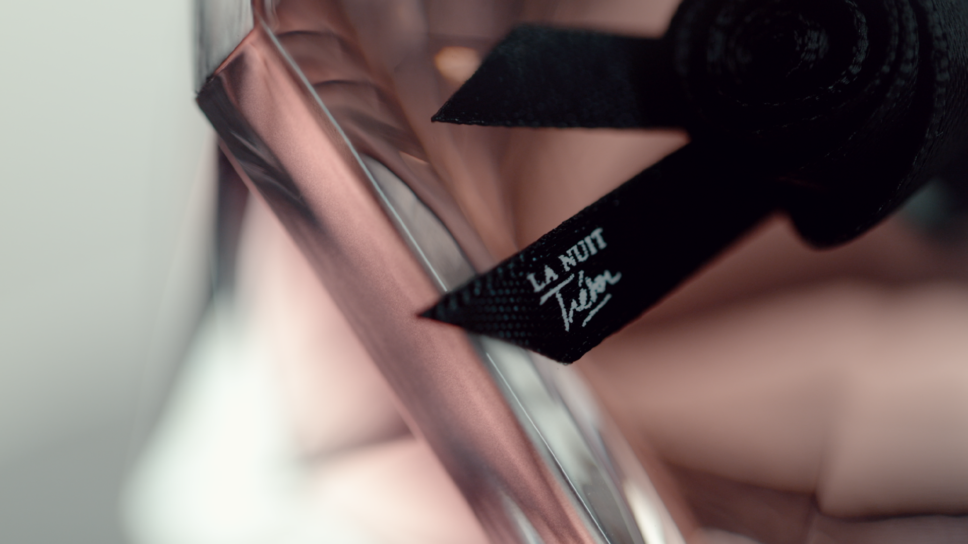 Close-up photo of a clothing tag that reads 'LA NUIT Tain' attached to a black fabric, with a shiny pink or metallic surface nearby.
