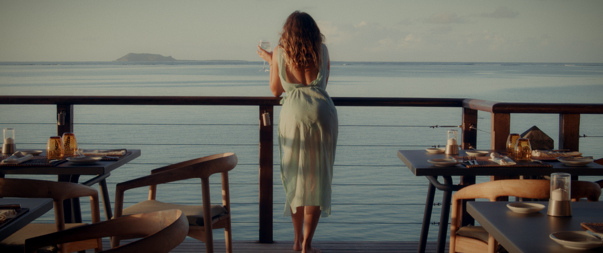 Woman standing barefoot on restaurant balcony overlooking ocean, holding a glass of wine, tables with tableware in foreground.