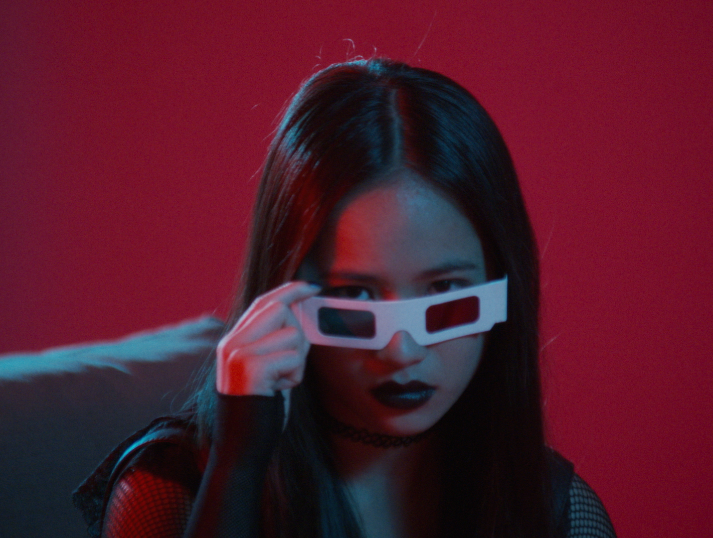 A woman with long dark hair holding 3D glasses, with a serious expression, against a red background.