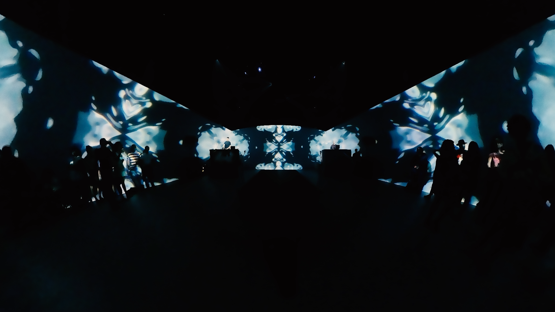 Dark room with large digital screens displaying abstract, symmetrical blue and white visuals. Silhouettes of people are standing and watching the screens, with some DJs or performers at the center.