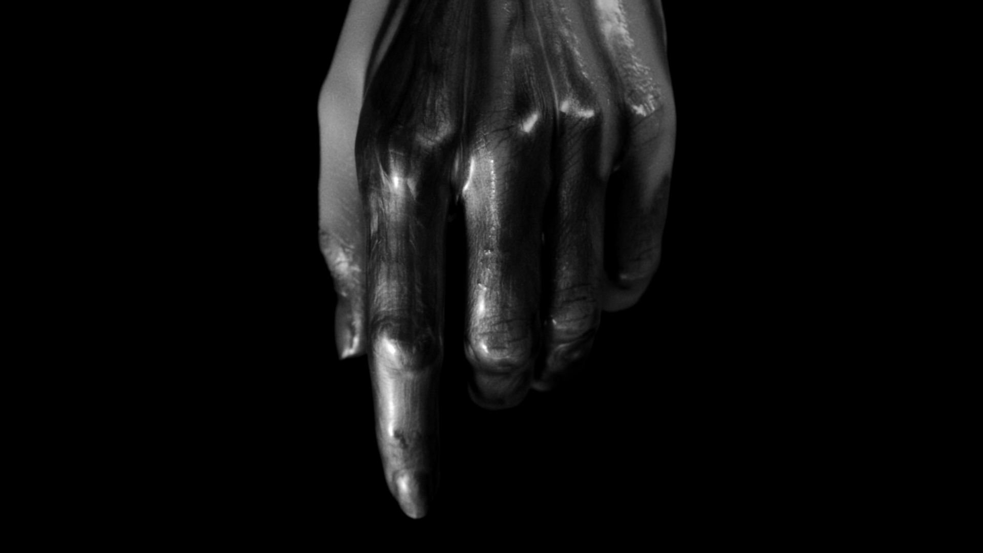 Black and white close-up photograph of a hand with long fingers, with the index finger extended downward, against a black background.