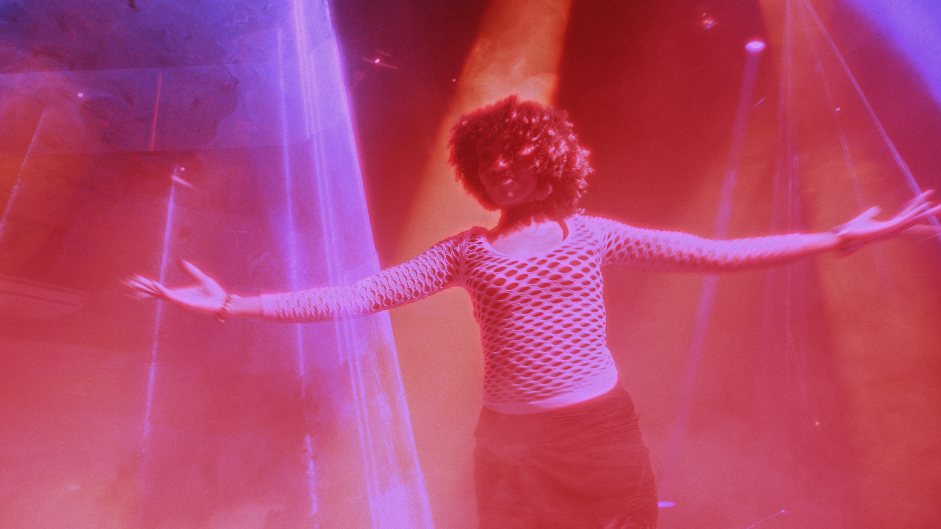 A woman with curly hair wearing a white fishnet top and a skirt stands with arms outstretched in a colorful, neon-lit environment with pink, purple, and red lighting.