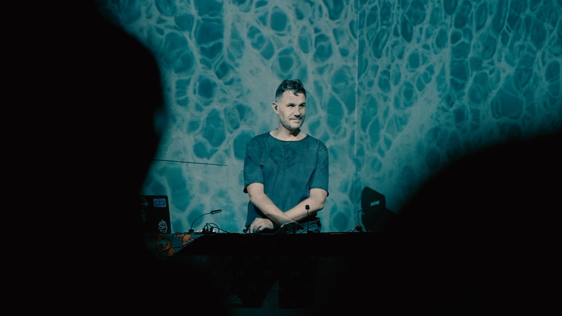 A man stands behind a DJ setup during a performance, with a blue textured background projected on the wall behind him.