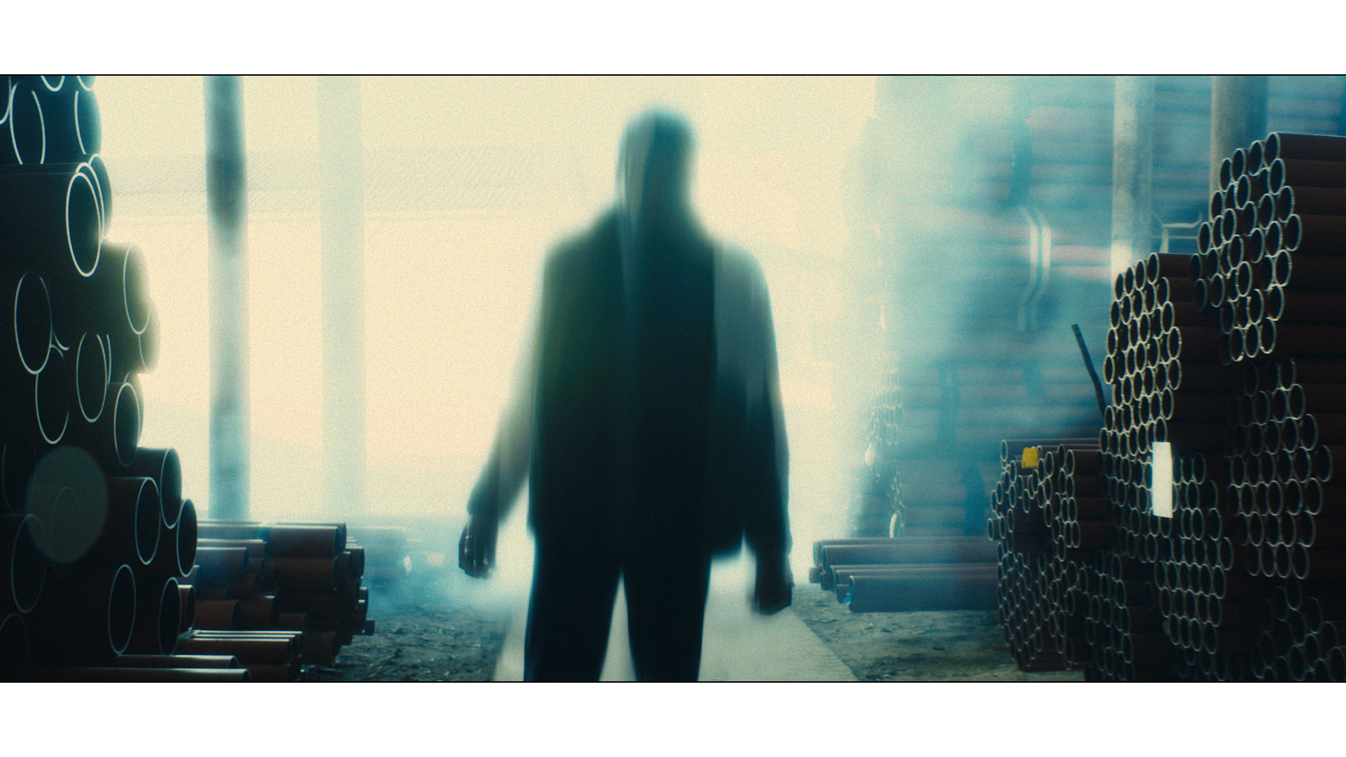 Silhouette of a person walking through an industrial warehouse with stacked pipes on both sides, dim lighting, and a misty or smoky atmosphere.