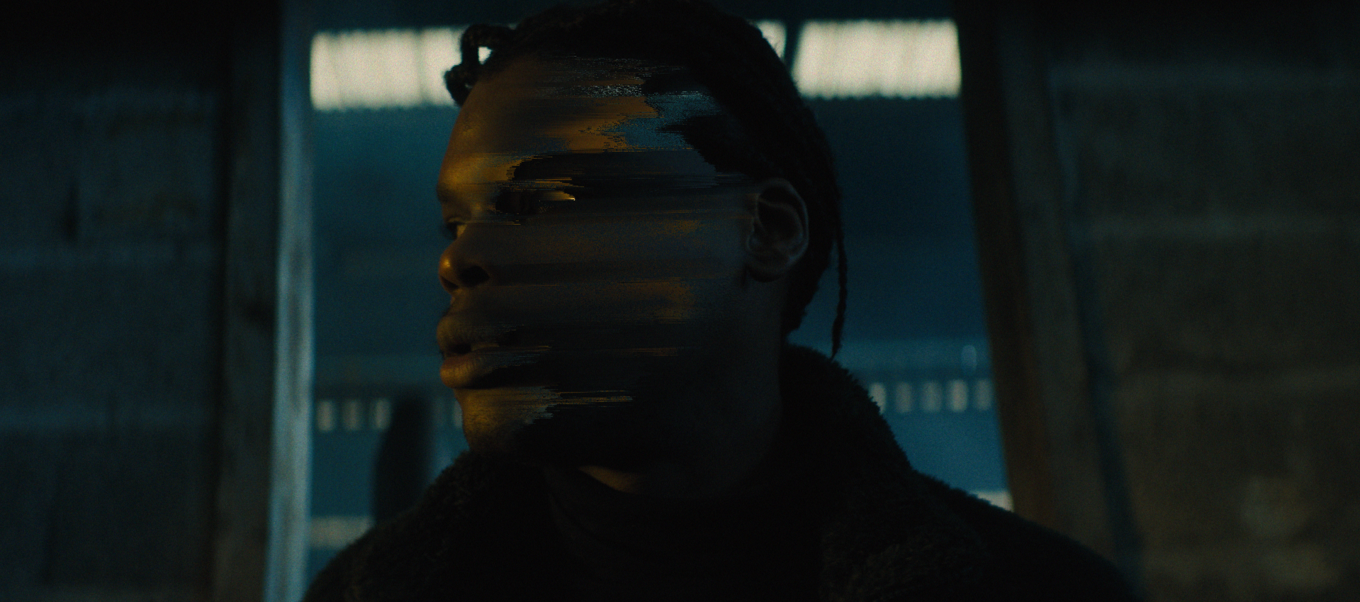 A woman with dark braided hair looking to the side in a dark, industrial setting with a window behind her.