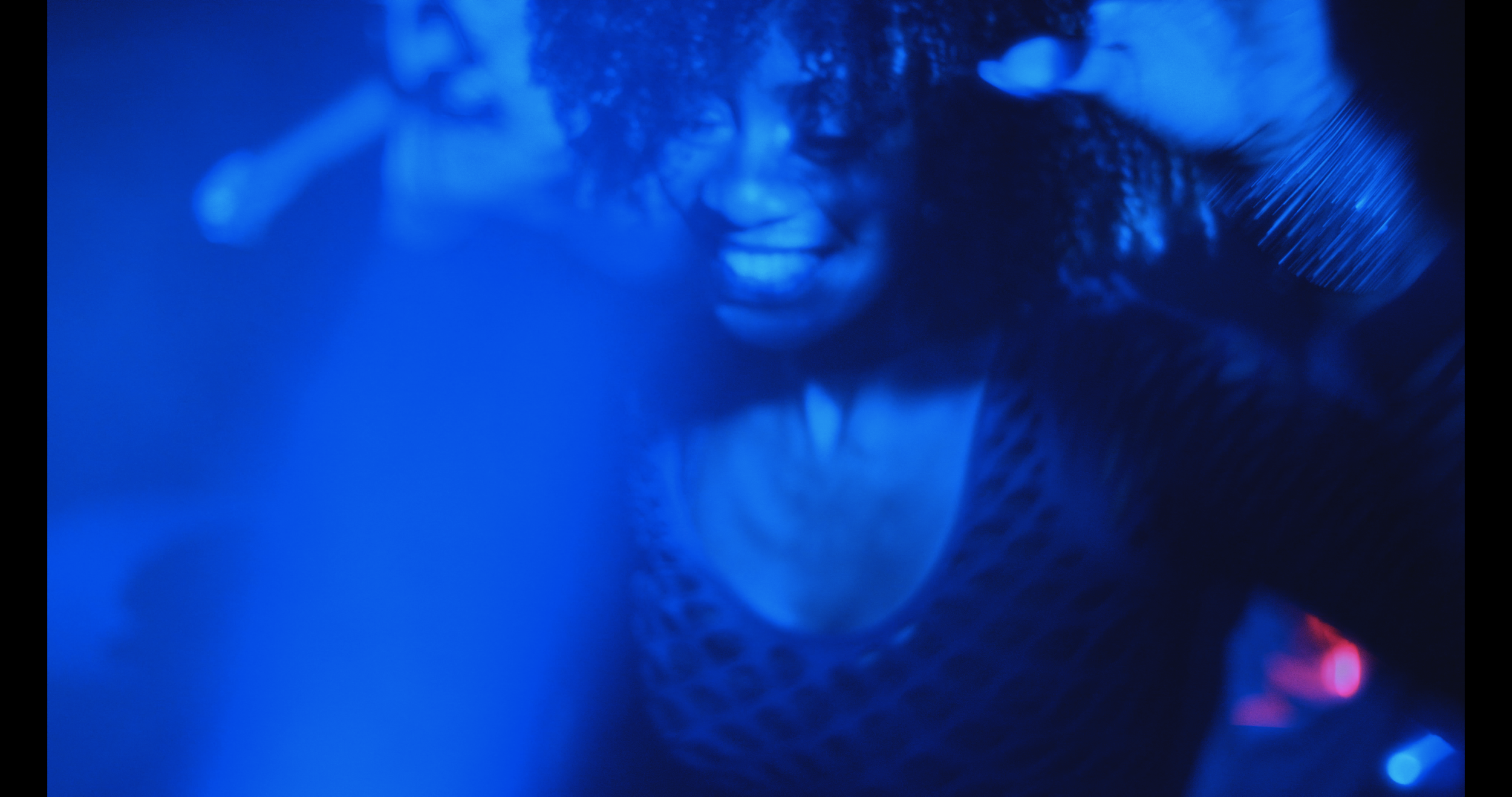 A woman smiling and reaching out with her hand, illuminated in blue lighting, with curly hair and wearing a patterned top.