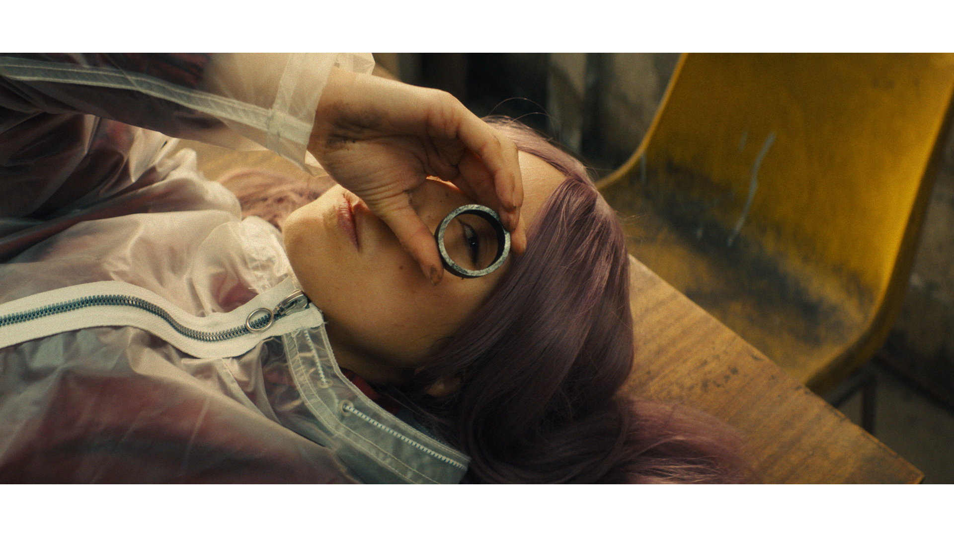 A woman with purple hair lying on a wooden surface, holding a circular object up to her right eye, with a yellow chair in the background.