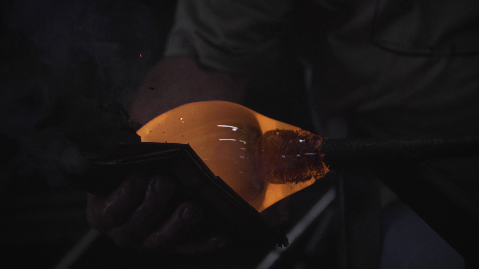 A person holding a glowing, heated glass object on a dark work surface, with tools and protective gloves visible.