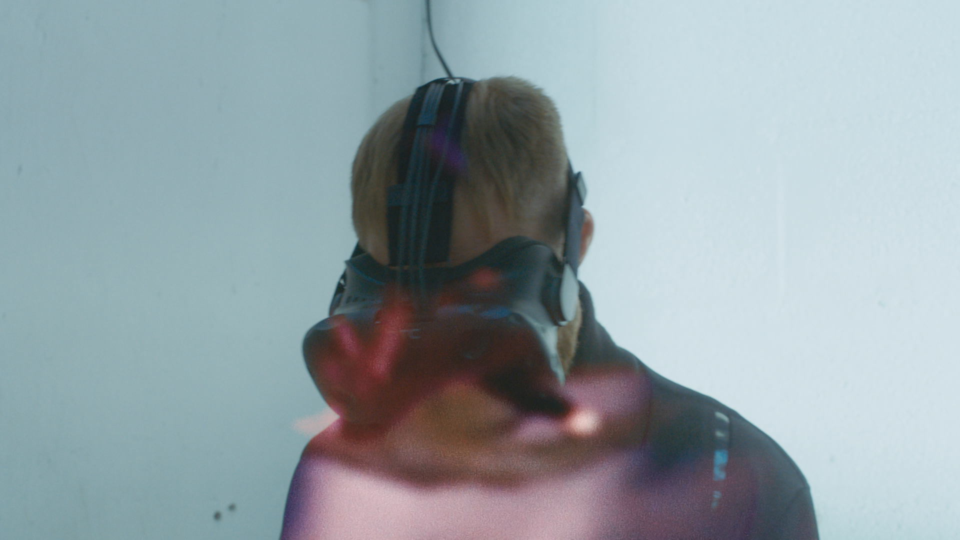 Back of a person with short hair wearing large headphones and a dark jacket, standing against a plain white wall.