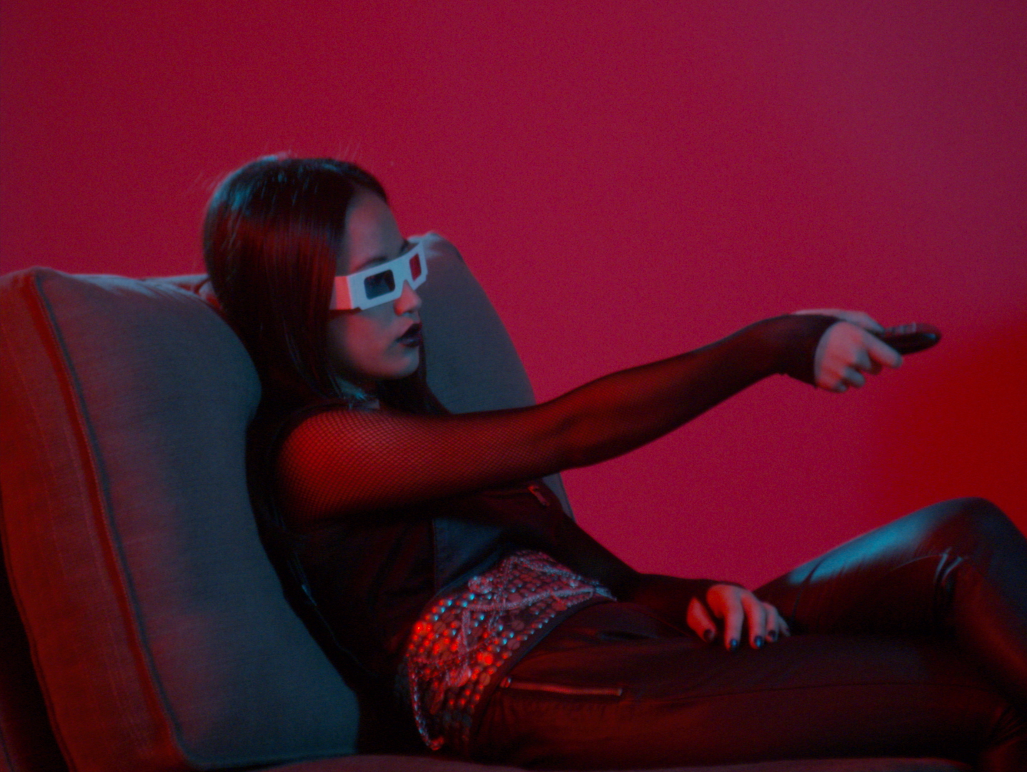A woman with long dark hair wearing 3D glasses, a black mesh top, and leather pants, reclining on a sofa and pointing a remote control, against a red background.