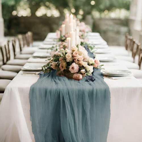 A long dining table set for a formal event, decorated with a blue table runner, pink and white floral arrangement, and white candles in the center.