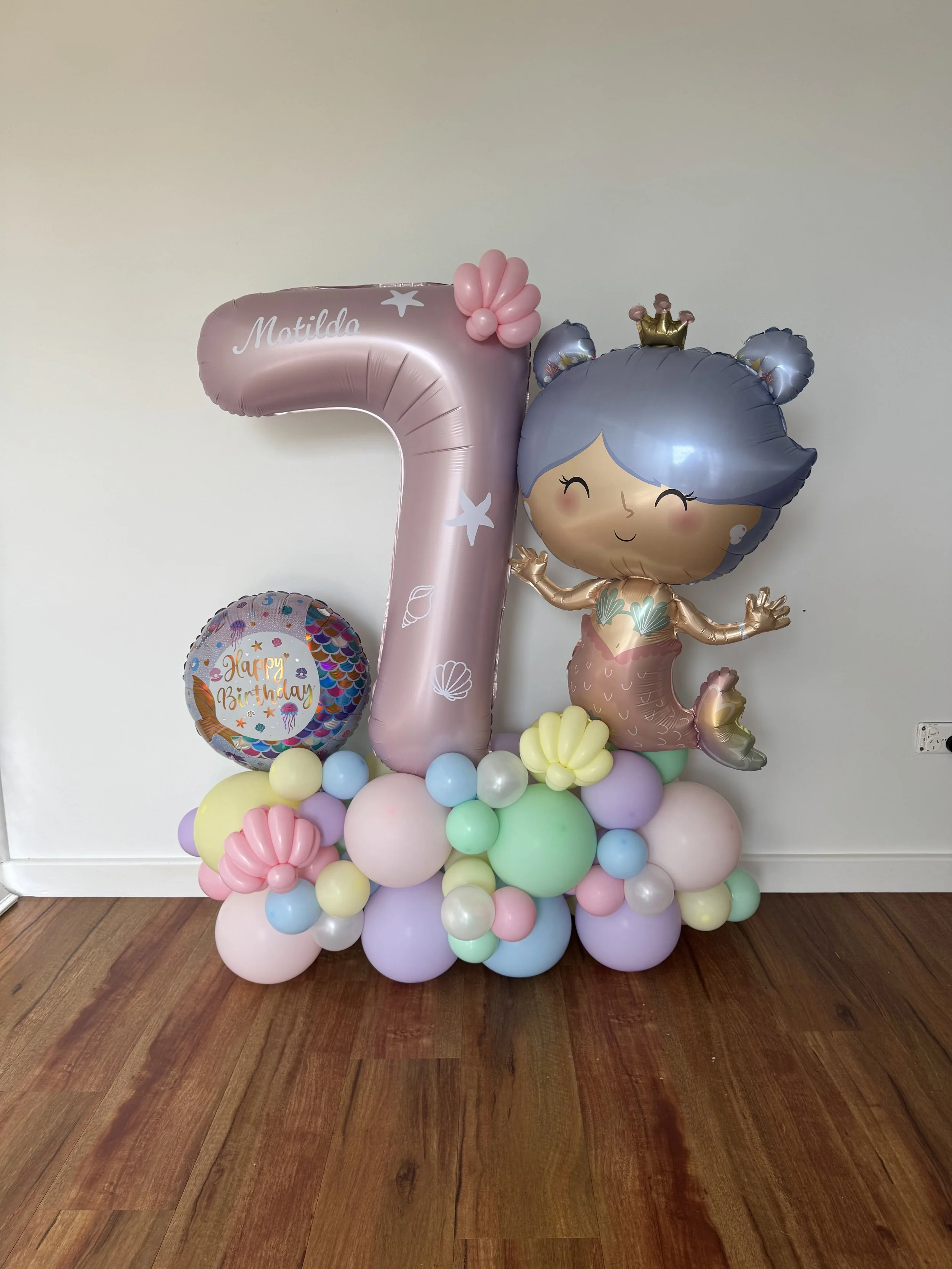Decorative balloon arrangement for a birthday, featuring a large pink number one balloon with  a mermaid character balloon, a round balloon with 'Happy Birthday' text, and clusters of pastel-colored balloons at the base on a wooden floor against a plain wall.