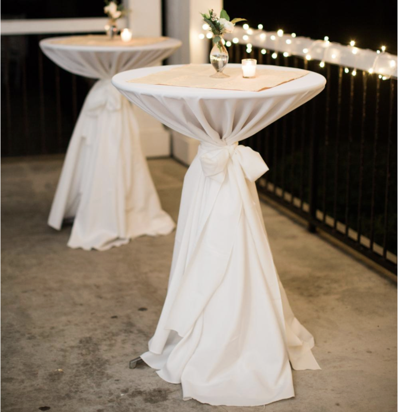 Two high-top tables covered with white cloths tied in the middle, decorated with a small flower vase and candles, set for an outdoor evening event with string lights in the background.