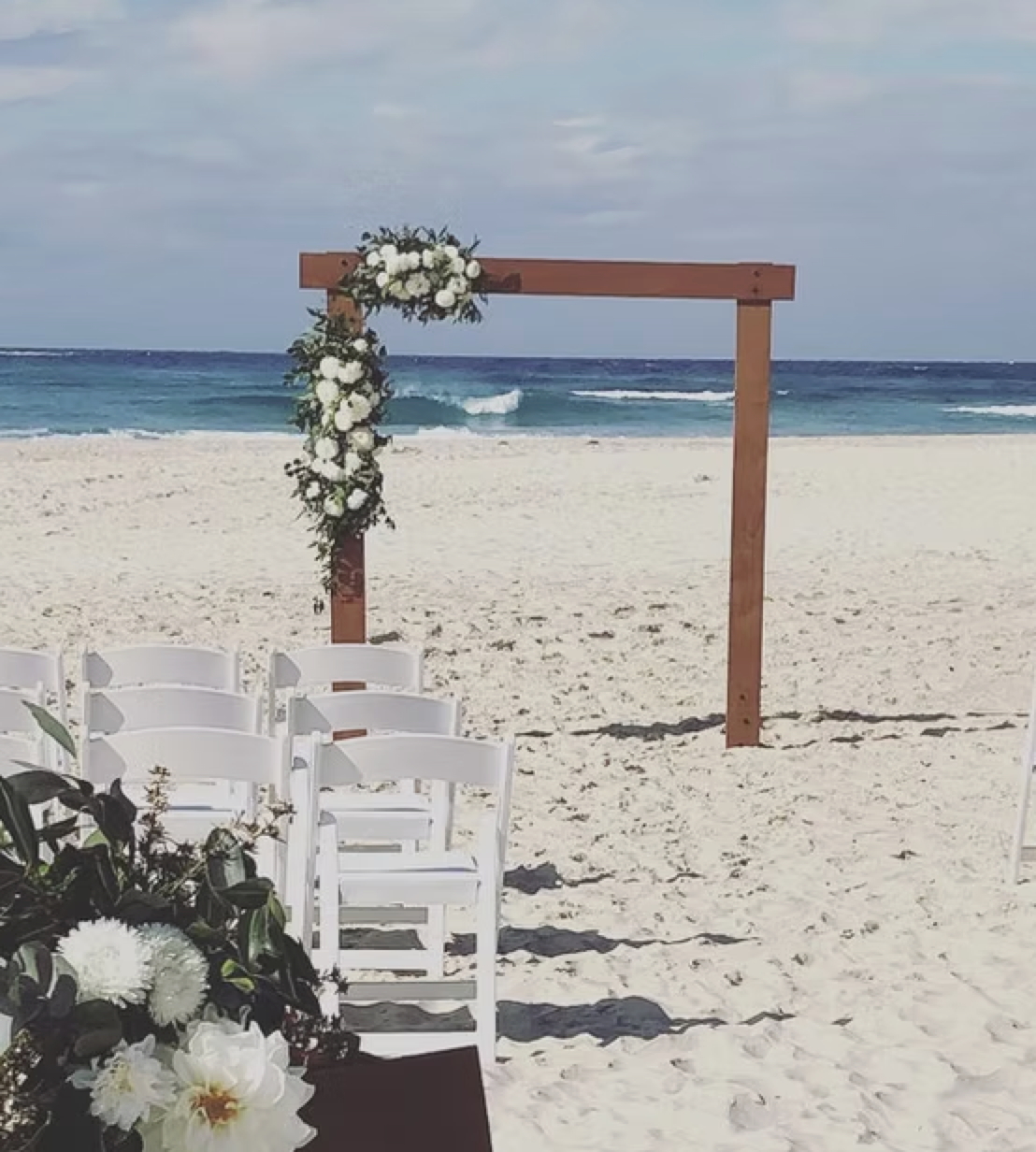 A wedding arch decorated with white flowers on a beach with white chairs and the ocean in the background.