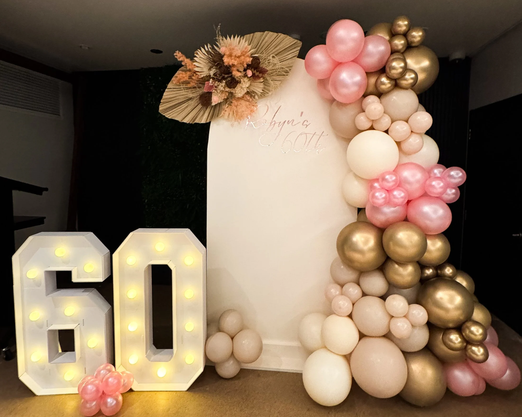 Decorative backdrop for a celebration, featuring a large illuminated '60' sign, a white panel with gold cursive writing, and a balloon garland with pink, white, and gold balloons, and a nearby flower arrangement.
