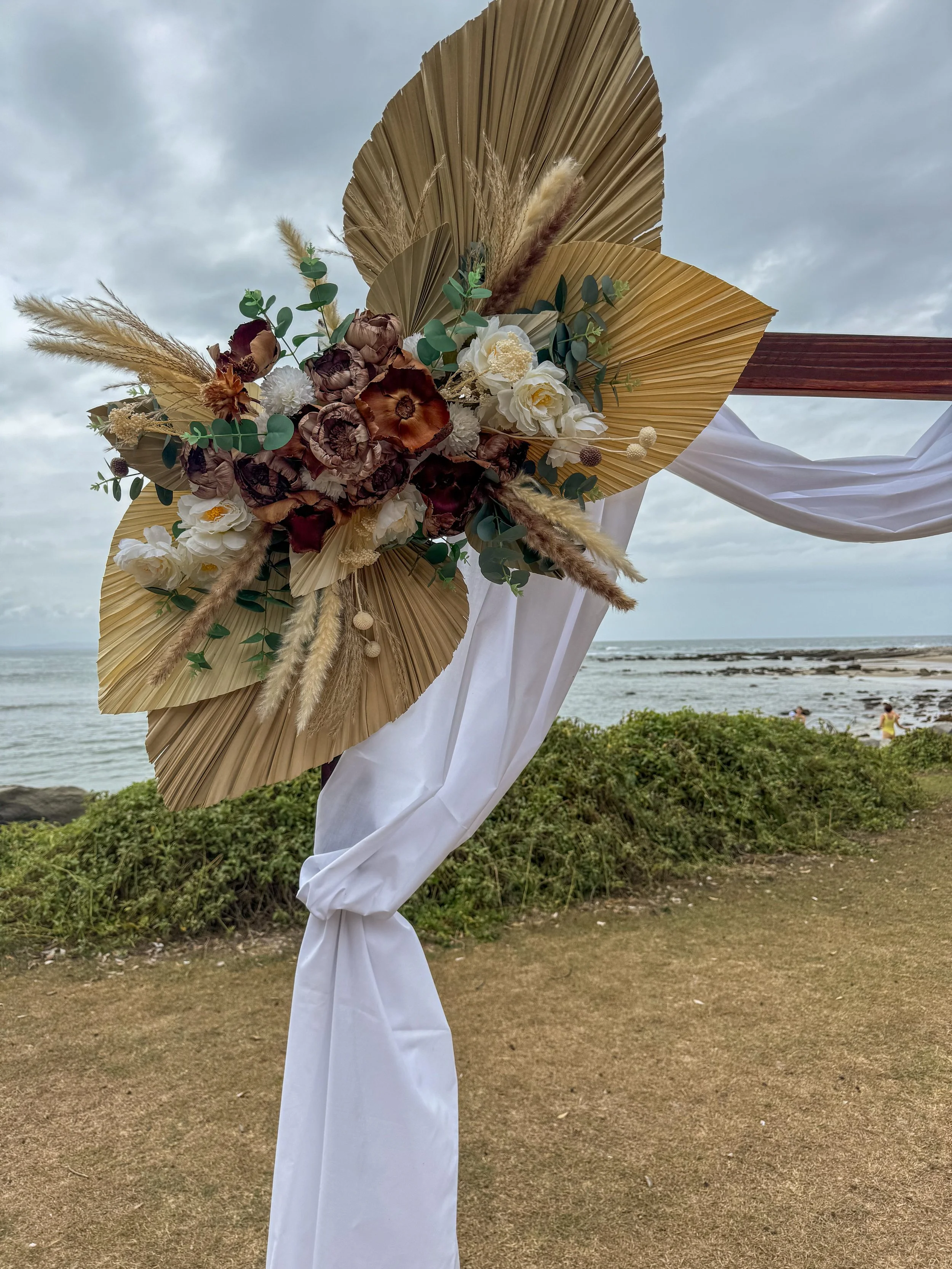 Decorative floral arrangement with dried and fresh flowers, eucalyptus, palm leaves, and pampas grass, attached to a wooden board decorated with white fabric, set outdoors near the beach with an overcast sky.