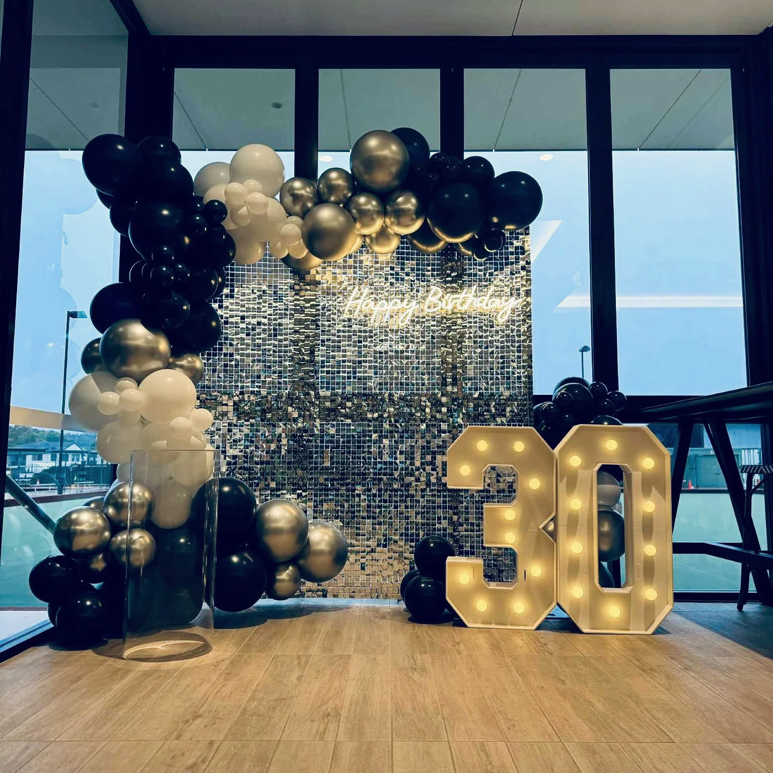 Birthday celebration setup with a large illuminated '30' in front, a backdrop made of small reflective tiles, a balloon arch in black, white, and metallic colors, and a neon sign that says 'Happy Birthday'.