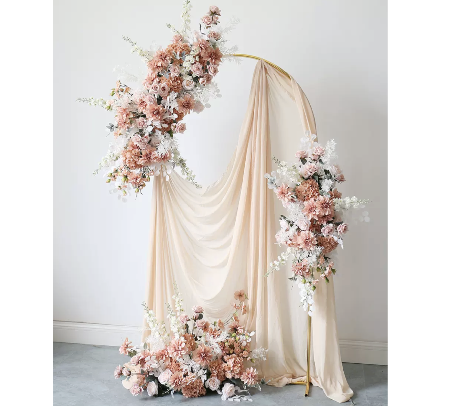 A wedding arch decorated with pink, peach, white, and cream flowers alongside flowing beige and cream drapes, set against a plain white wall.
