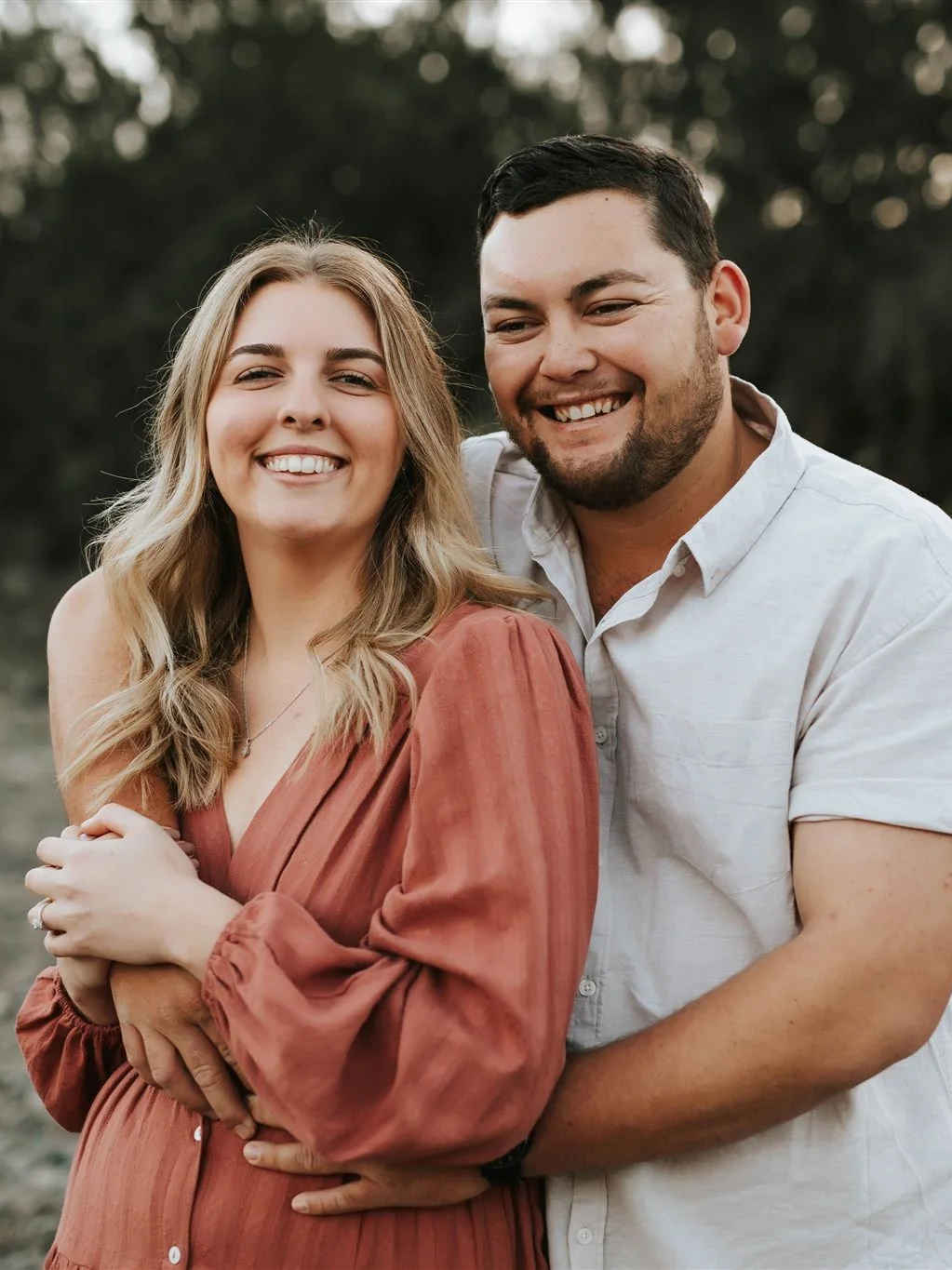 A smiling couple poses outdoors with greenery in the background, the woman has blonde hair and is wearing a rust-colored dress, and the man has dark hair and a beard, wearing a white shirt.
