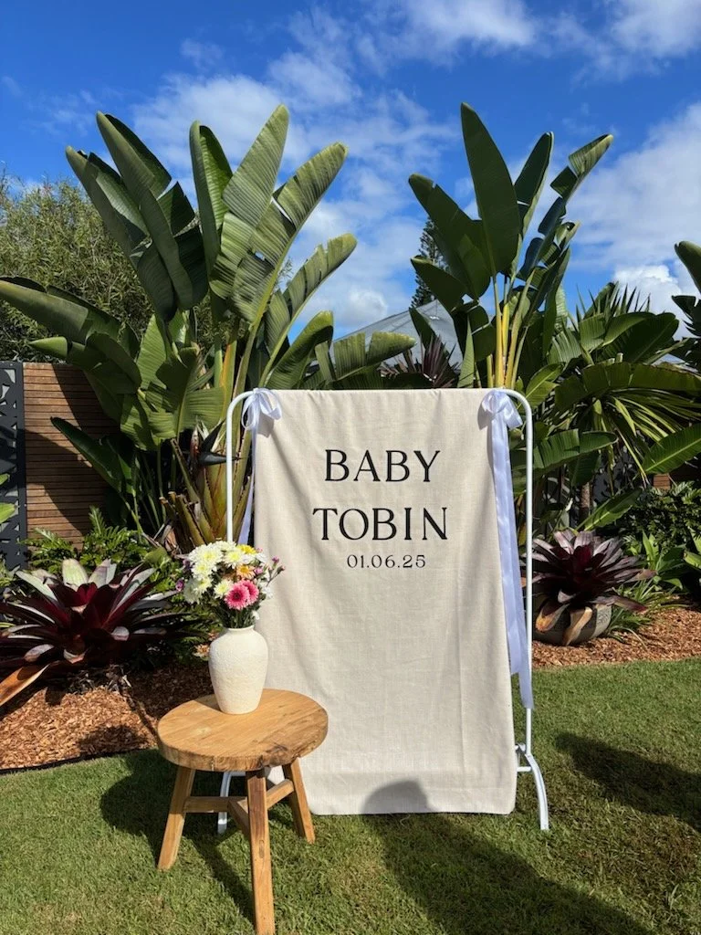 Outdoor setup with a white fabric banner marked "Baby Tobin" with the date "01.06.25," supported by a white frame. A small wooden stool with a white vase of colorful flowers is placed in front. Behind are large lush tropical plants and a partly cloudy blue sky.