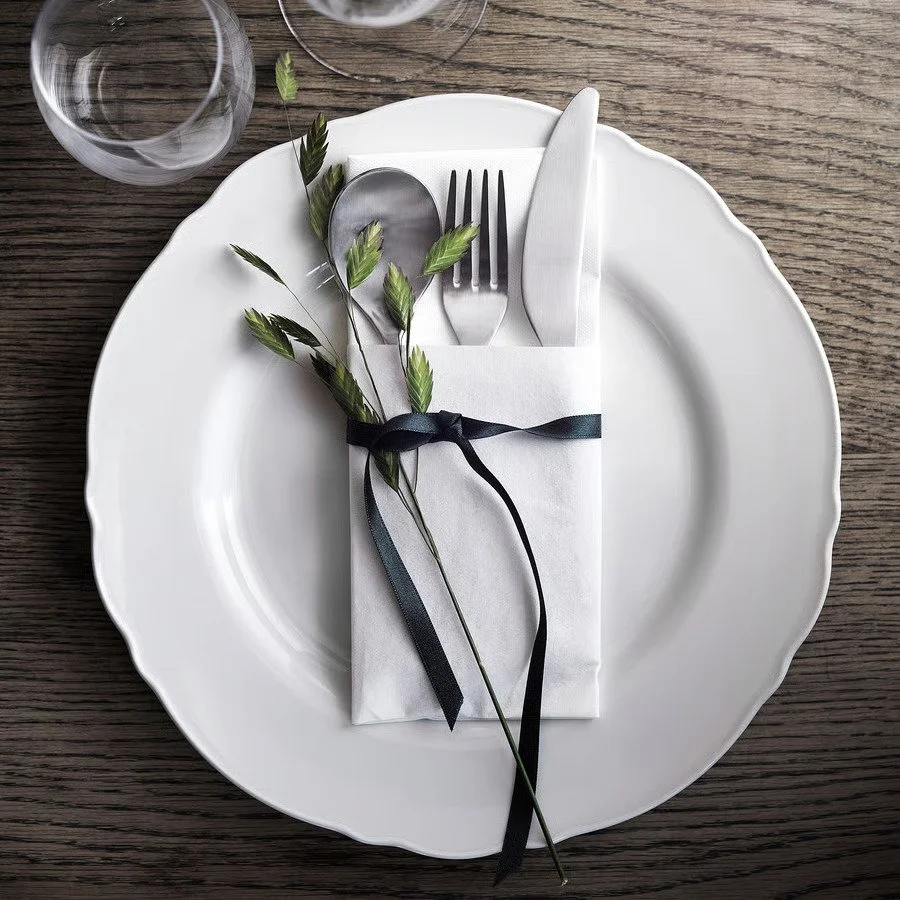 A white plate with a napkin-wrapped set of silverware, decorated with green foliage and tied with a black ribbon, on a wooden table.