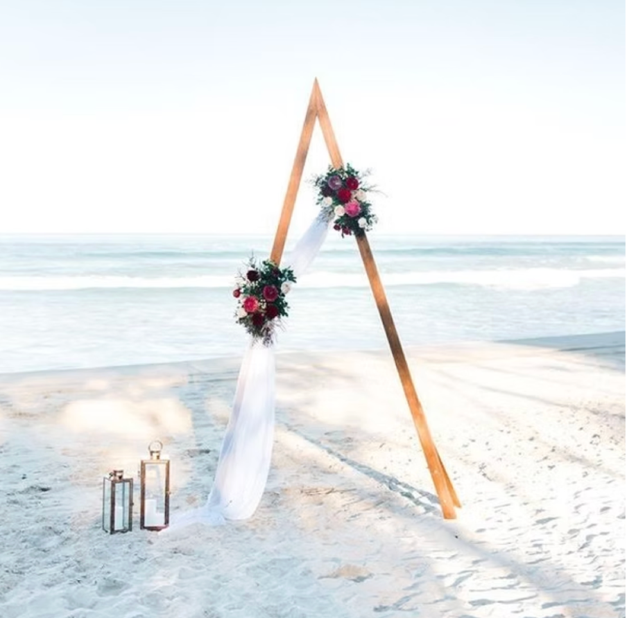 Wedding arch decorated with flowers and white fabric on a beach, with lanterns nearby.