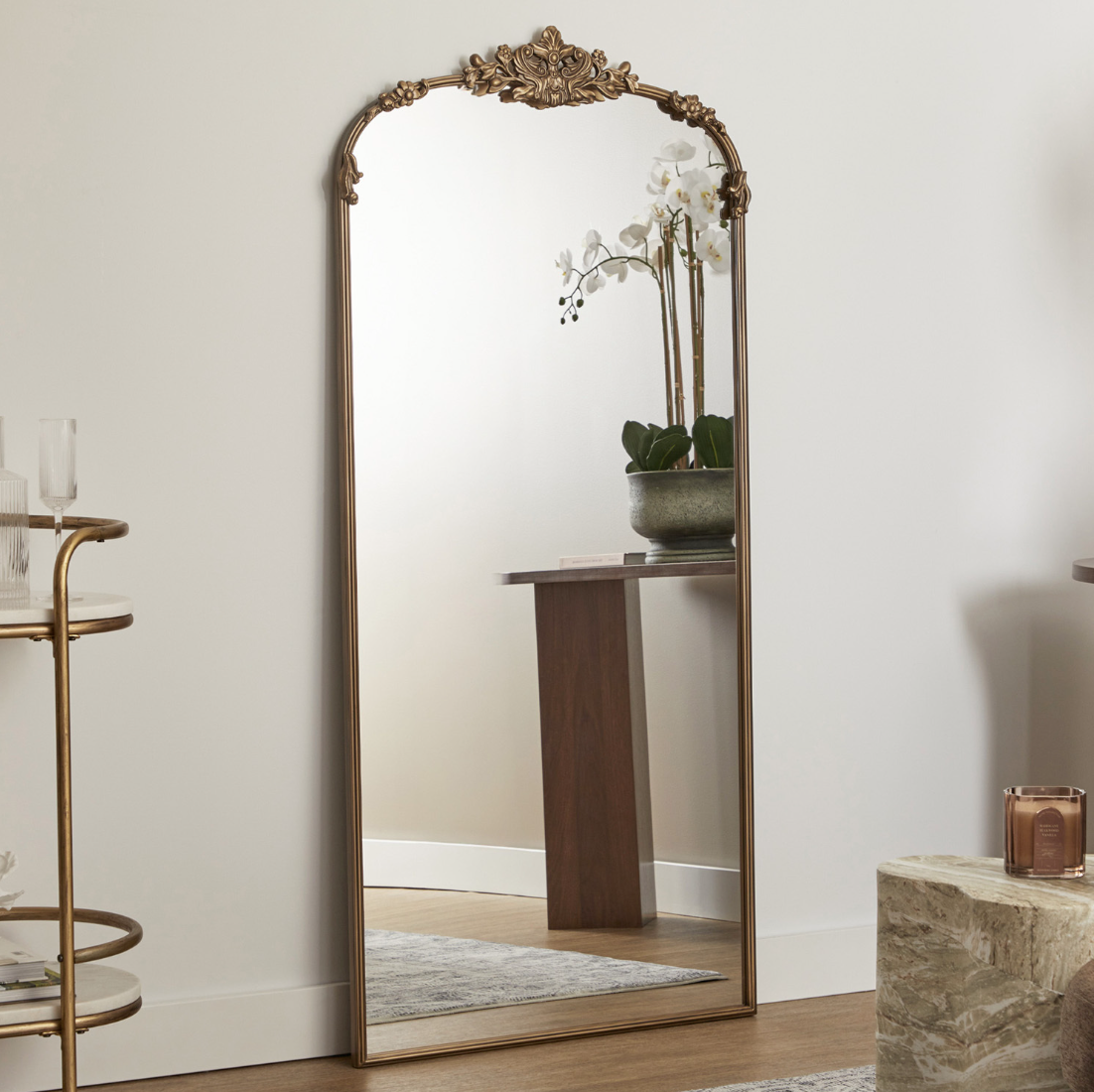 A tall, ornate, oval mirror with a gold-colored frame leaning against a white wall, reflecting a flower arrangement in a gray pot on a wooden table.