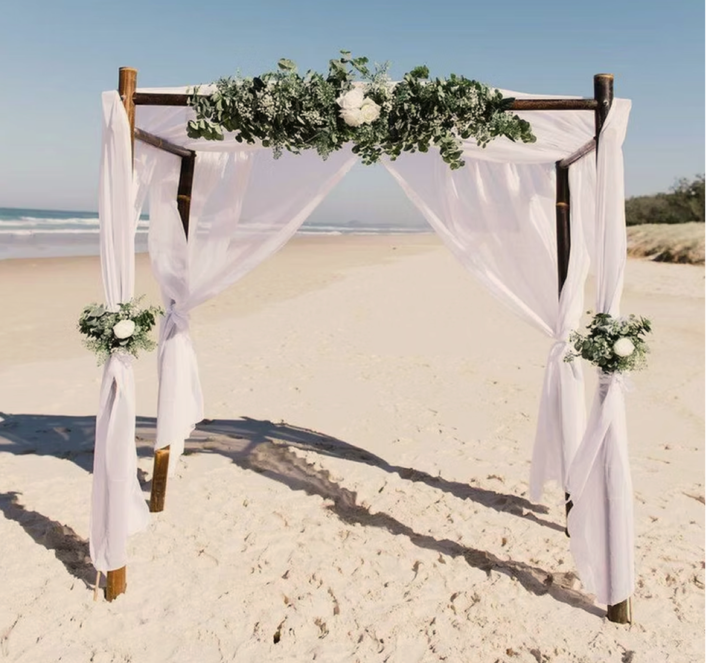 Wedding arch on a sandy beach decorated with white curtains and green floral arrangements.