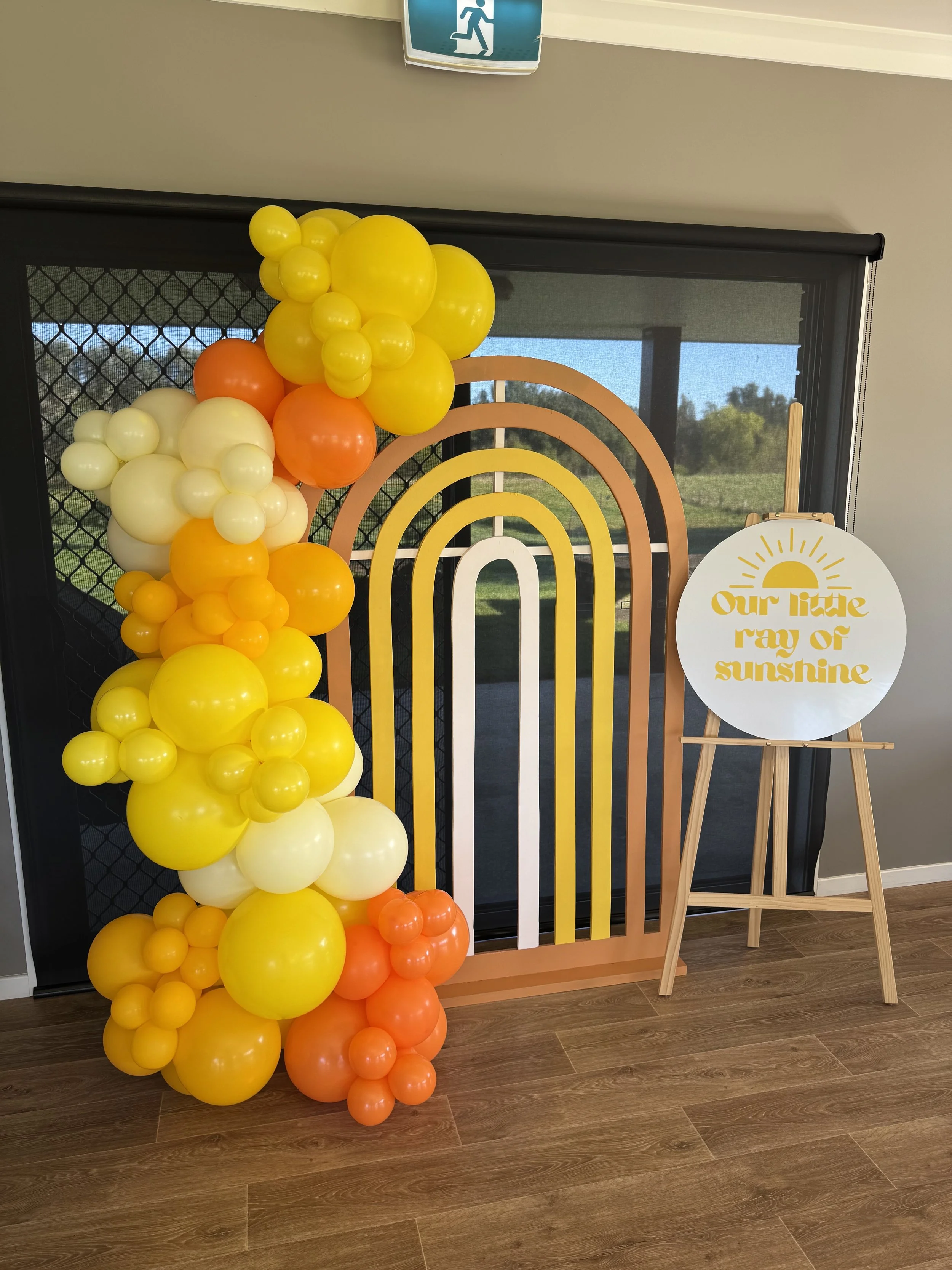 Decorative display with a colorful balloon arch in shades of yellow, orange, and white, a rainbow-themed backdrop, and a sign reading 'Our little ray of sunshine' on an easel.