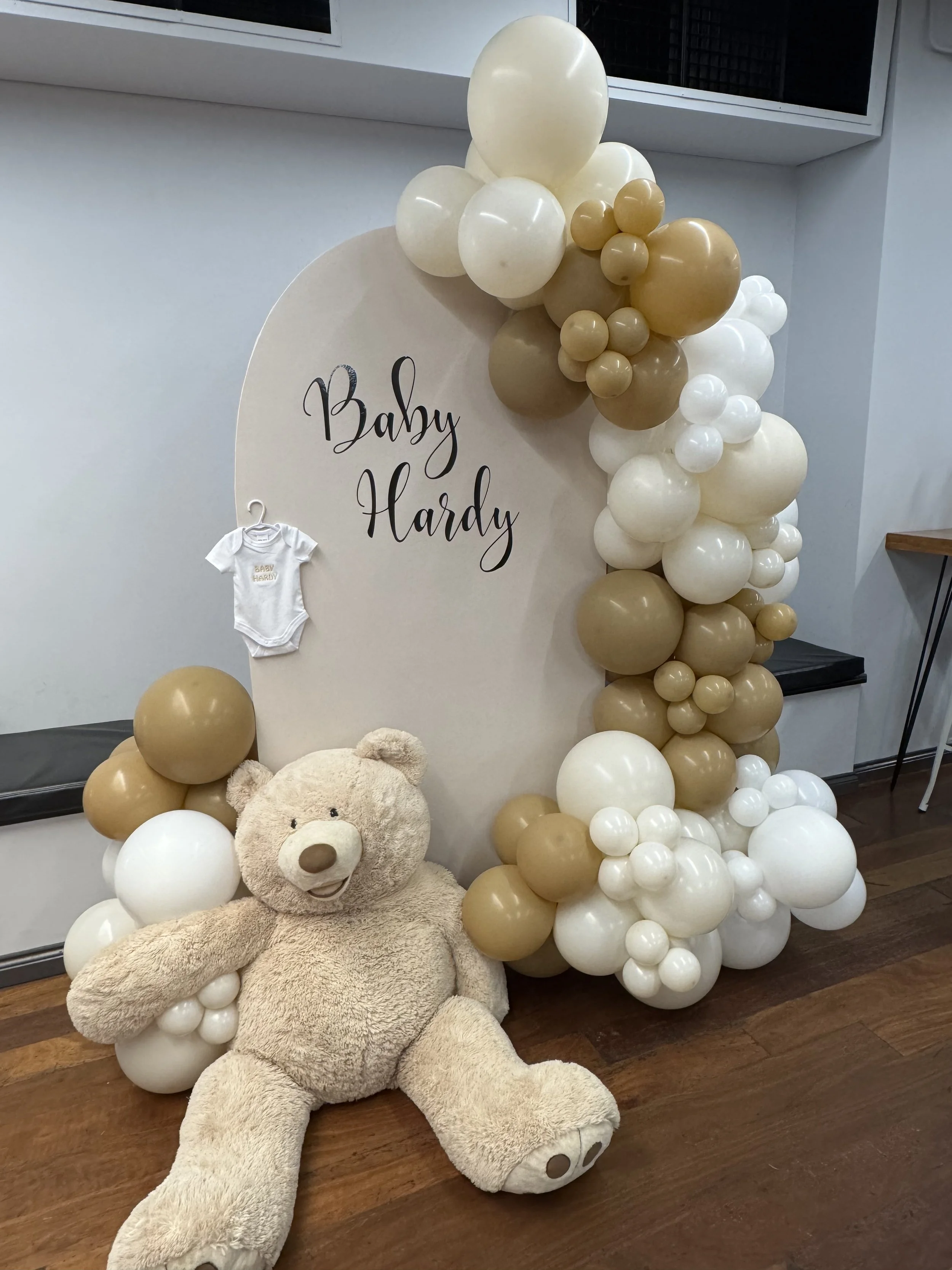 Decorative display for a baby shower with balloons in white, cream, and gold, a large teddy bear and an arch with custom writing.