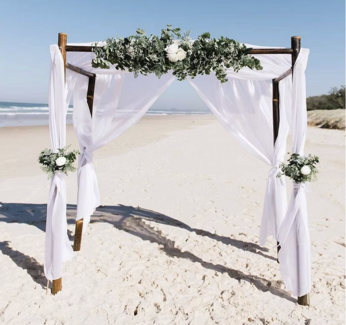 A beach wedding arch decorated with white fabric and green foliage with white flowers, on sandy beach with ocean and blue sky in the background.