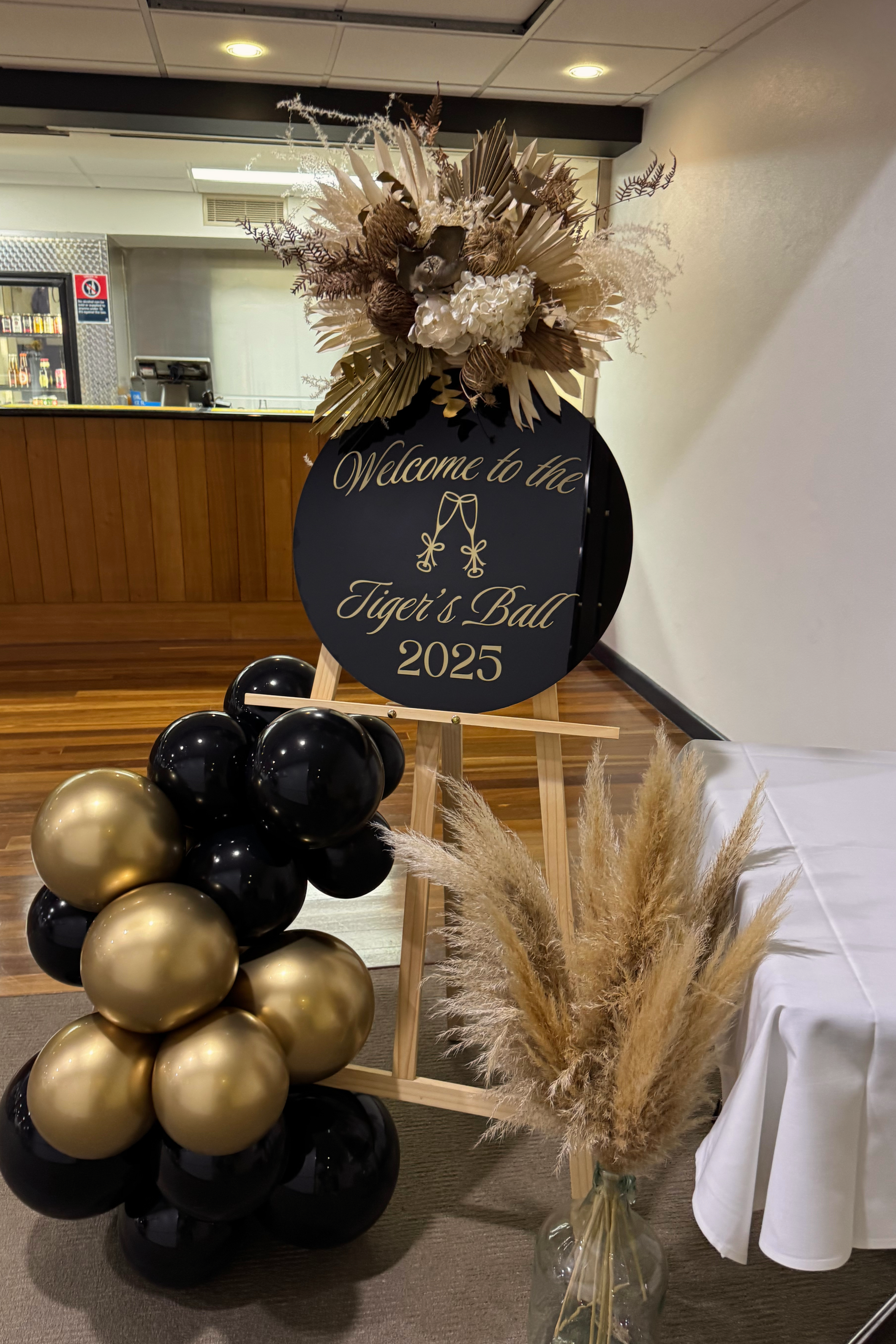 Decorative welcome sign for the Tiger's Ball 2025 with floral arrangement and black and gold balloons at an indoor event space.