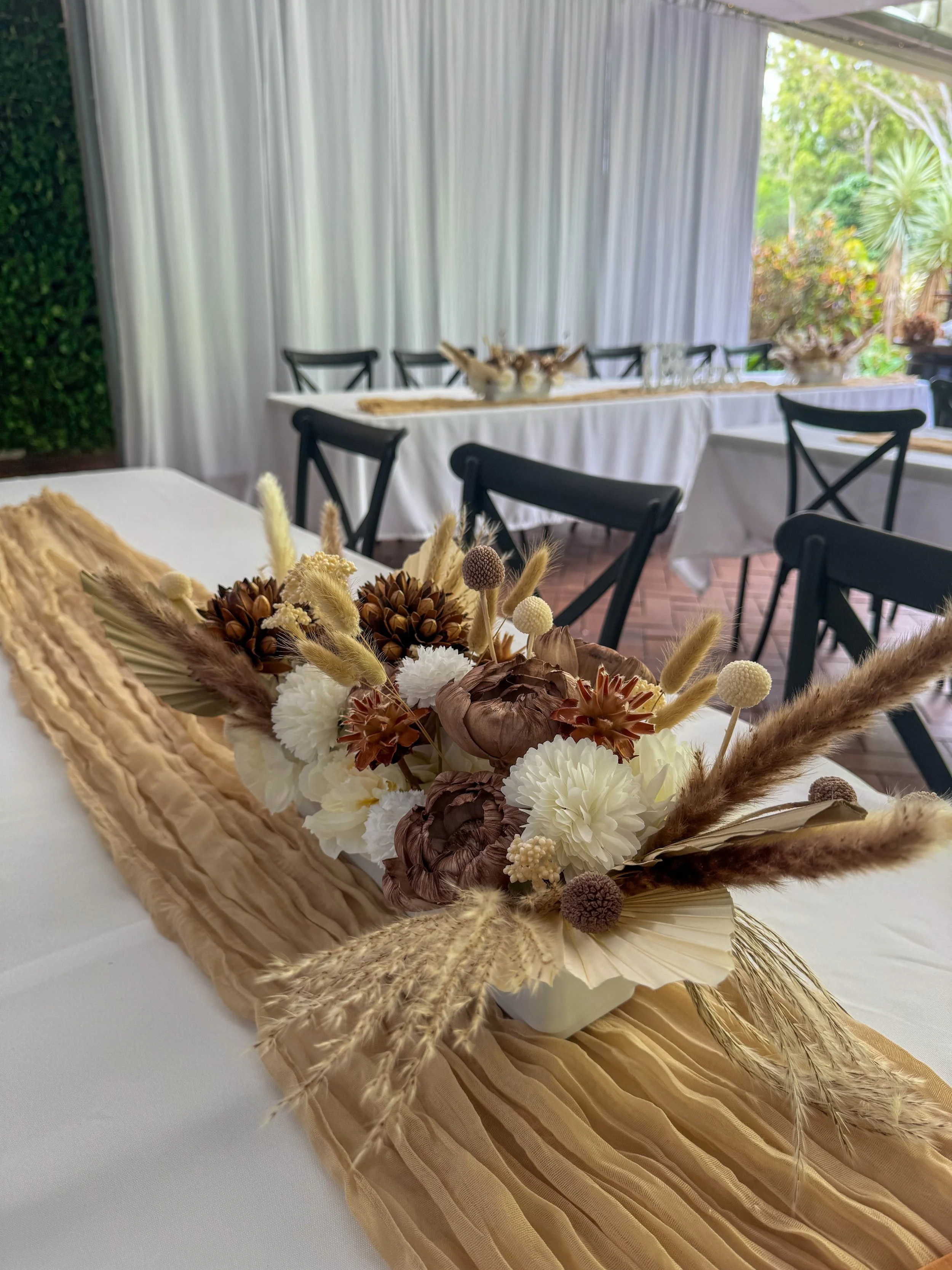 A neutral-toned floral centerpiece on a table with a beige ruffled table runner at an outdoor event, with additional tables and chairs in the background.