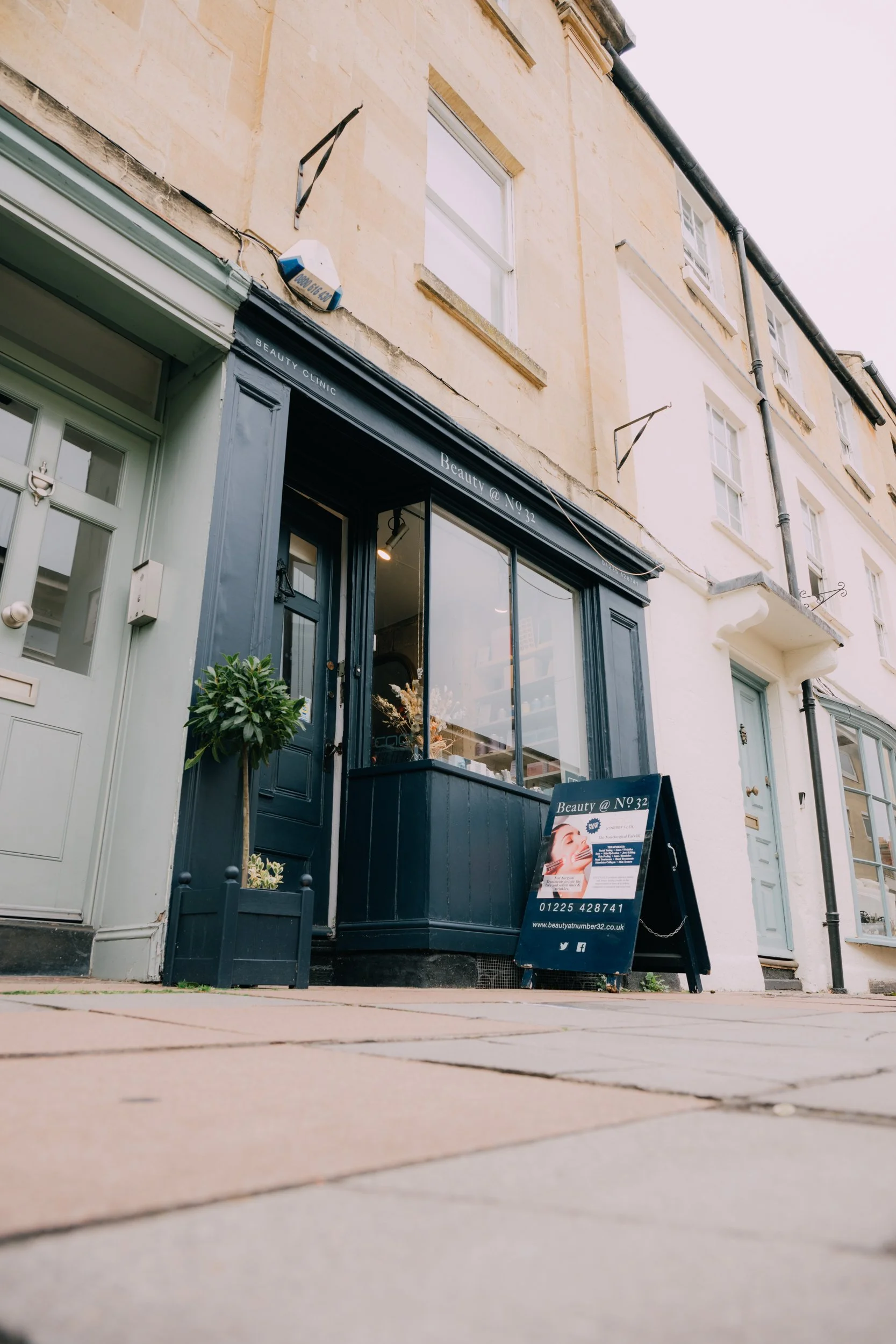 Outside view of a beauty clinic named 'Beauty @ No 32' with a dark blue entrance, large window, and a sidewalk sign displaying services and contact information, in front of cream-colored buildings with multiple floors and windows.