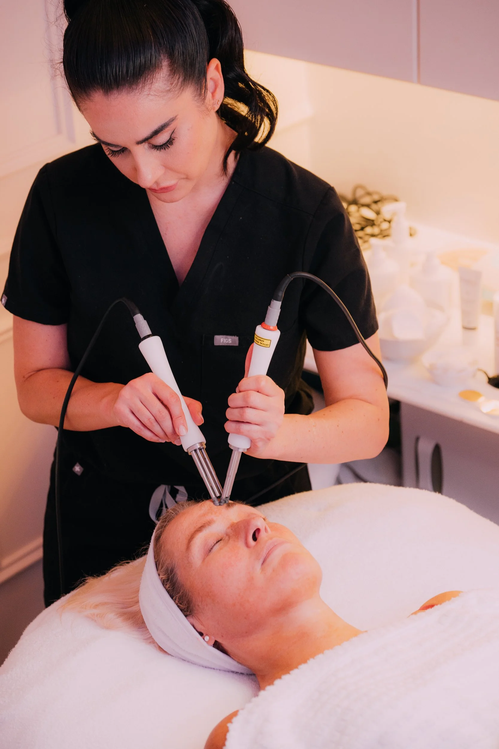 A woman lying on a treatment bed receiving a cosmetic facial procedure from a medical professional using two handheld devices.