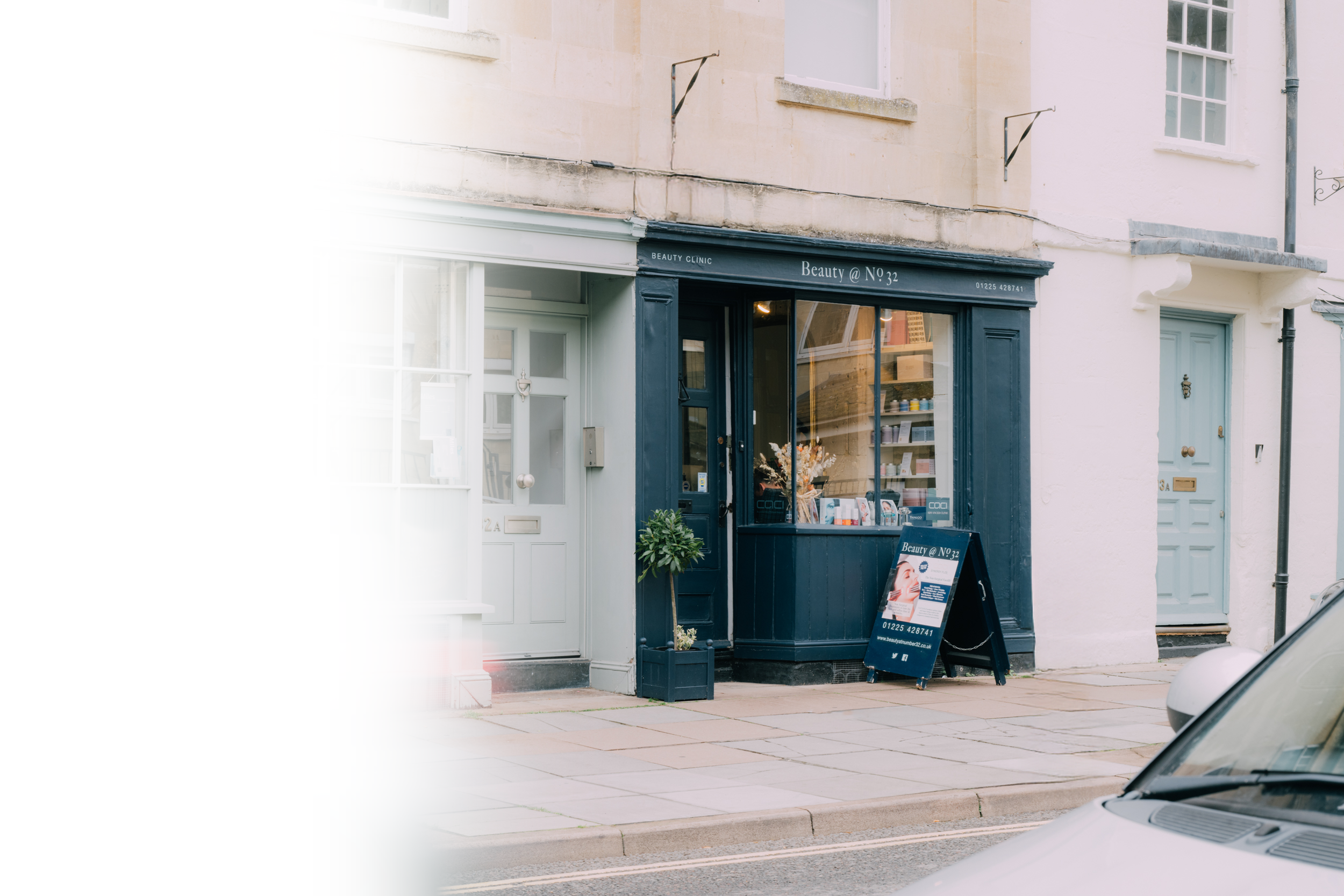 Small storefront called 'Beauty @ No 32' with a dark blue exterior, large window display, and a sandwich board sign outside on the sidewalk. The shop appears to offer beauty or skincare products.