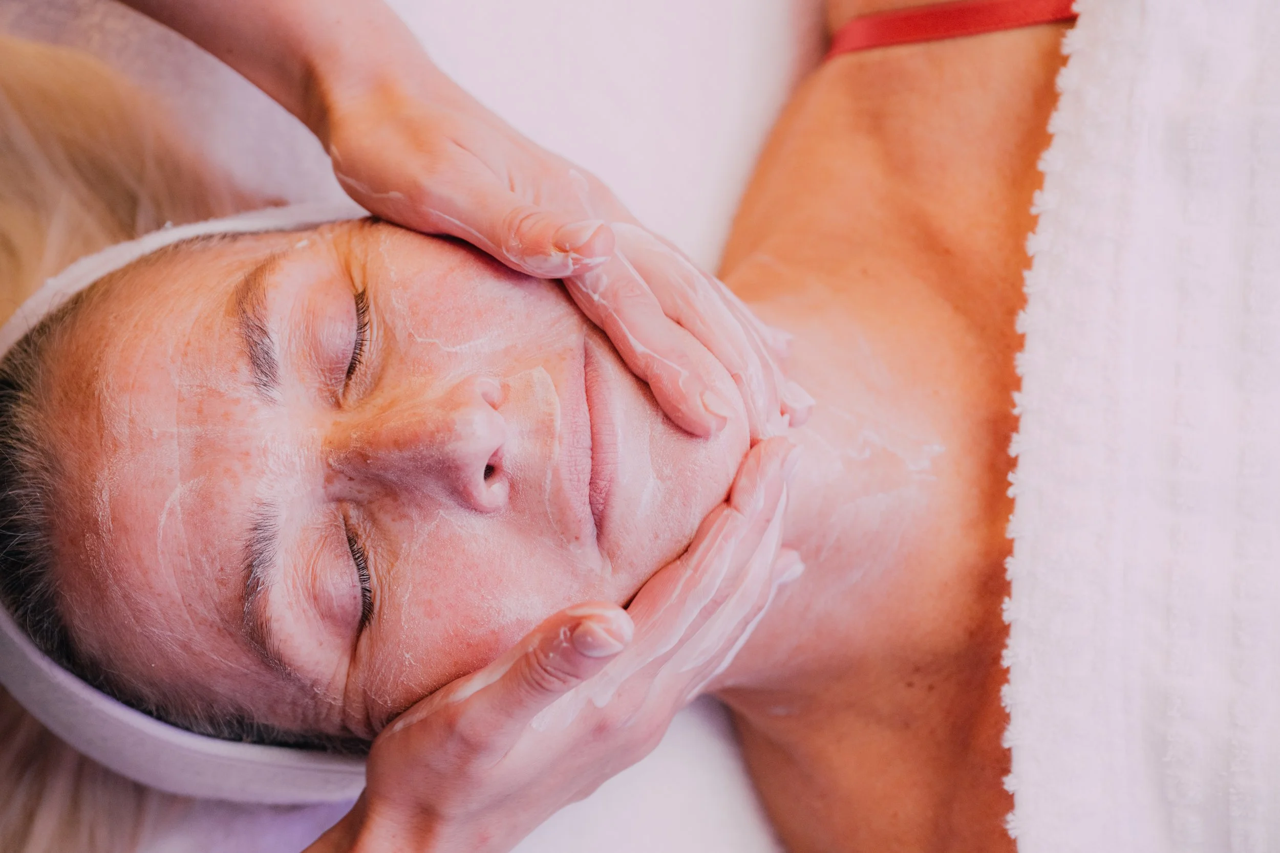 A person receiving a facial massage or skincare treatment, with their eyes closed and a towel covering their chest.
