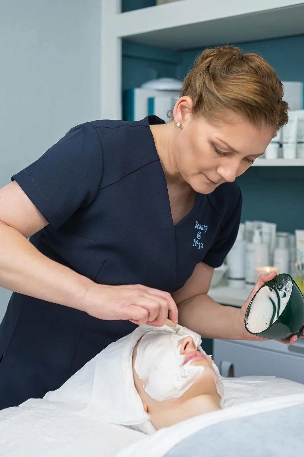A cosmetologist applying a facial mask to a woman lying on a treatment bed in a spa or skincare clinic.