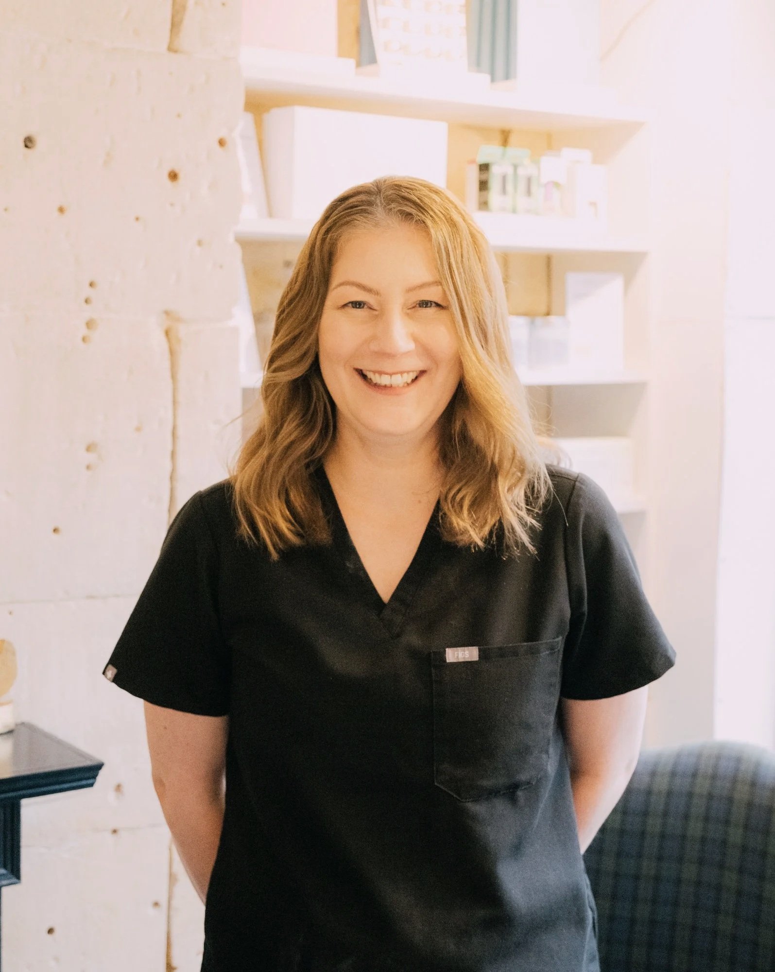 A woman with shoulder-length wavy blonde hair, smiling, wearing a black medical scrub top, standing indoors in front of white shelves with medical supplies.