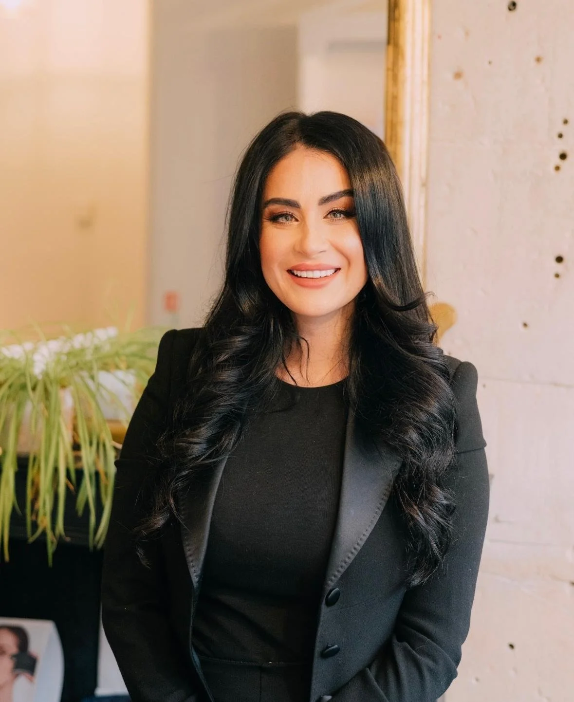 A smiling woman with long black hair wearing a black blazer and black top, standing indoors in front of a white brick wall and a wooden frame.