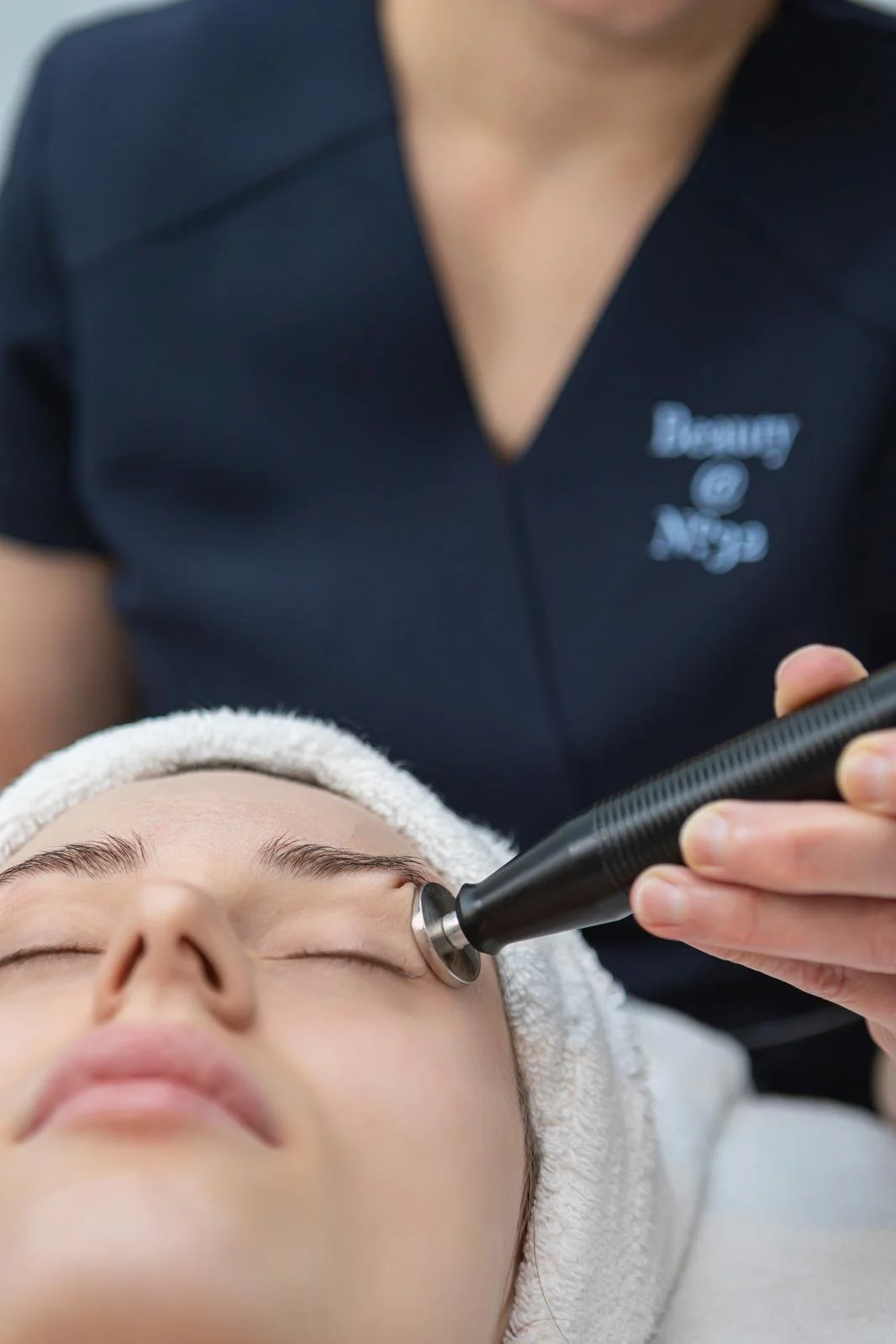A woman receives a facial treatment with a handheld skincare device from an professional wearing a navy blue uniform.