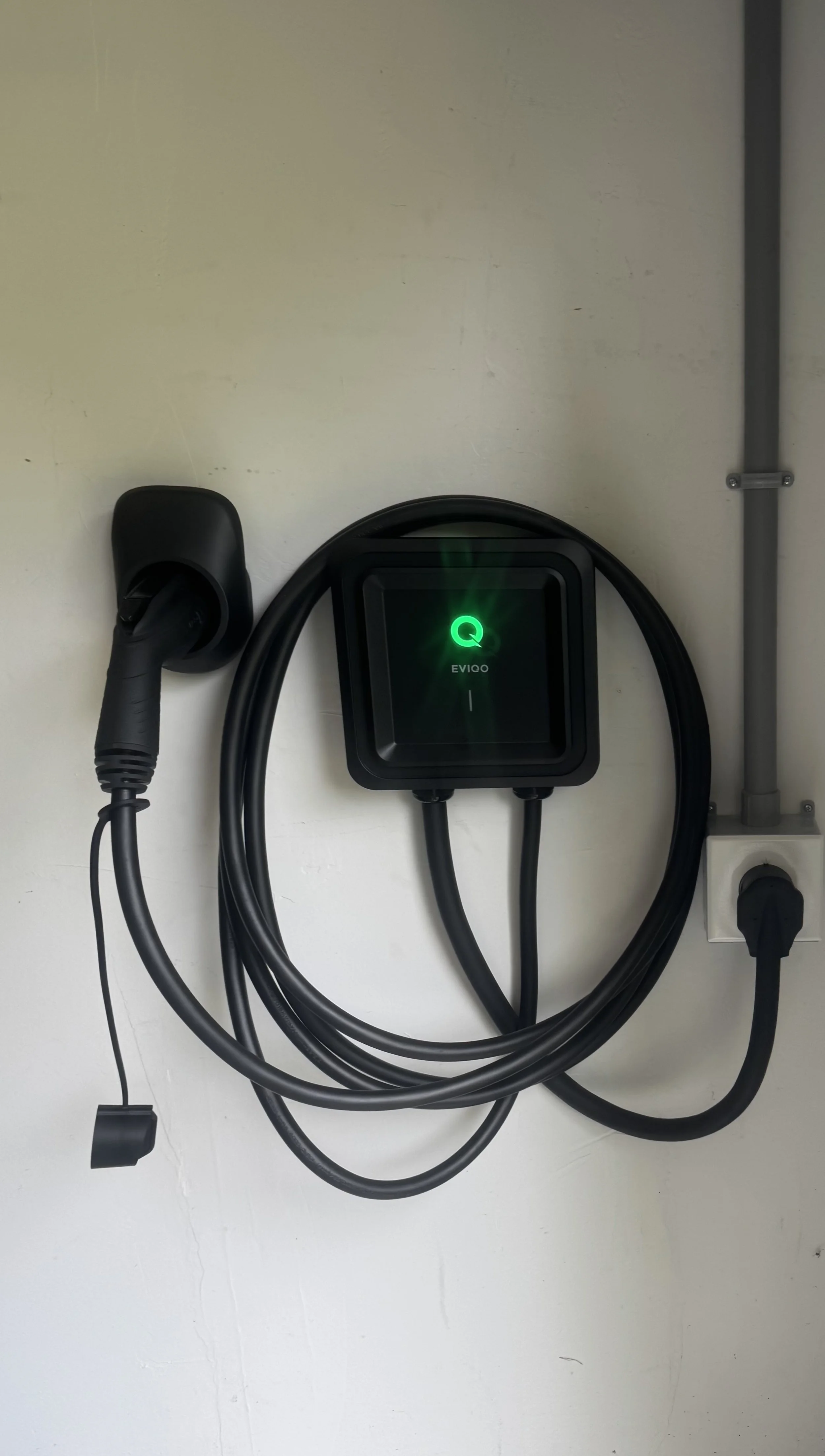 A wall-mounted electric vehicle charging station with a cable and plug, with a green light indicating it is ready for use.