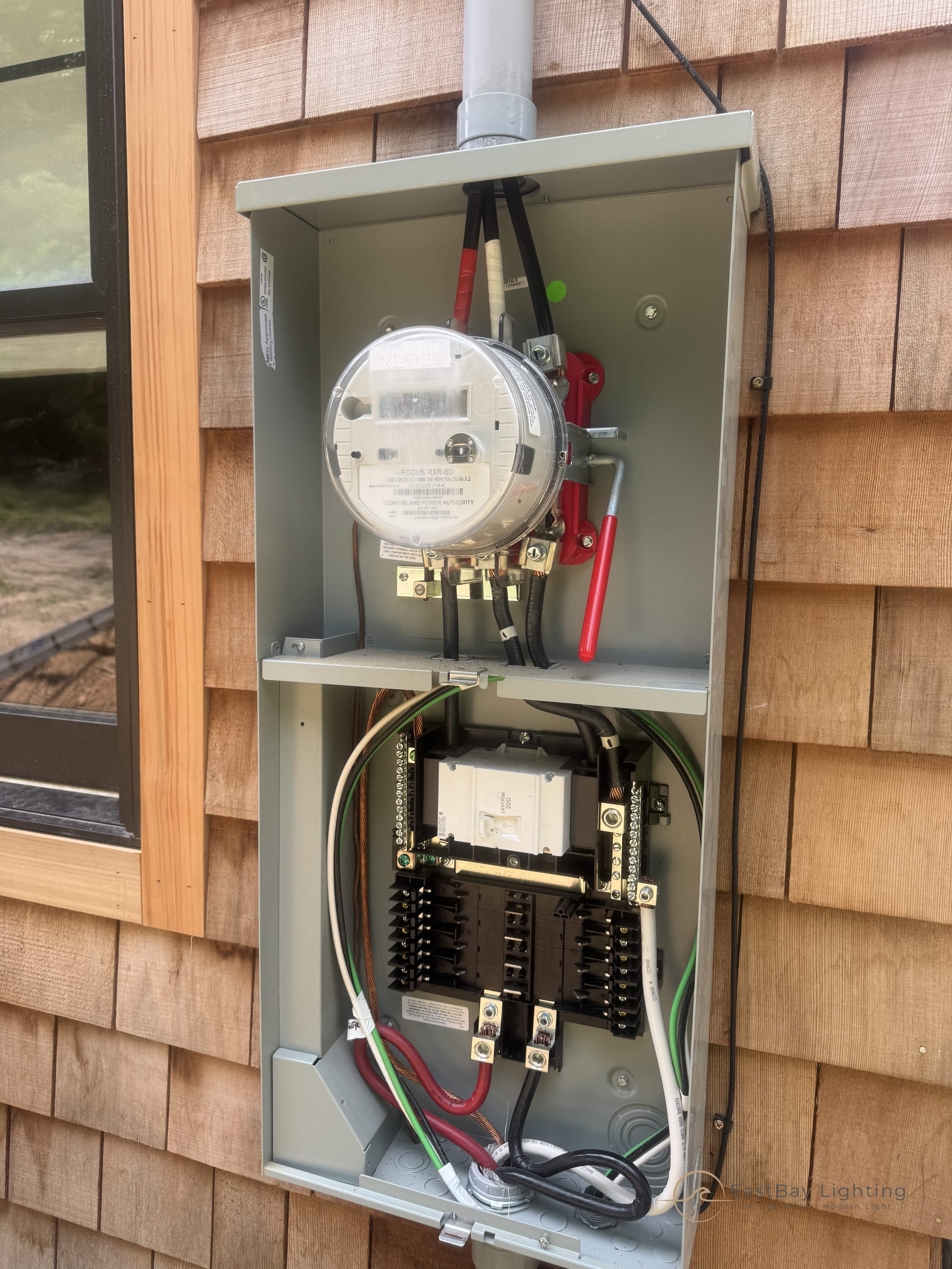 Open electrical meter box on the side of a wooden house showing wiring and components inside.