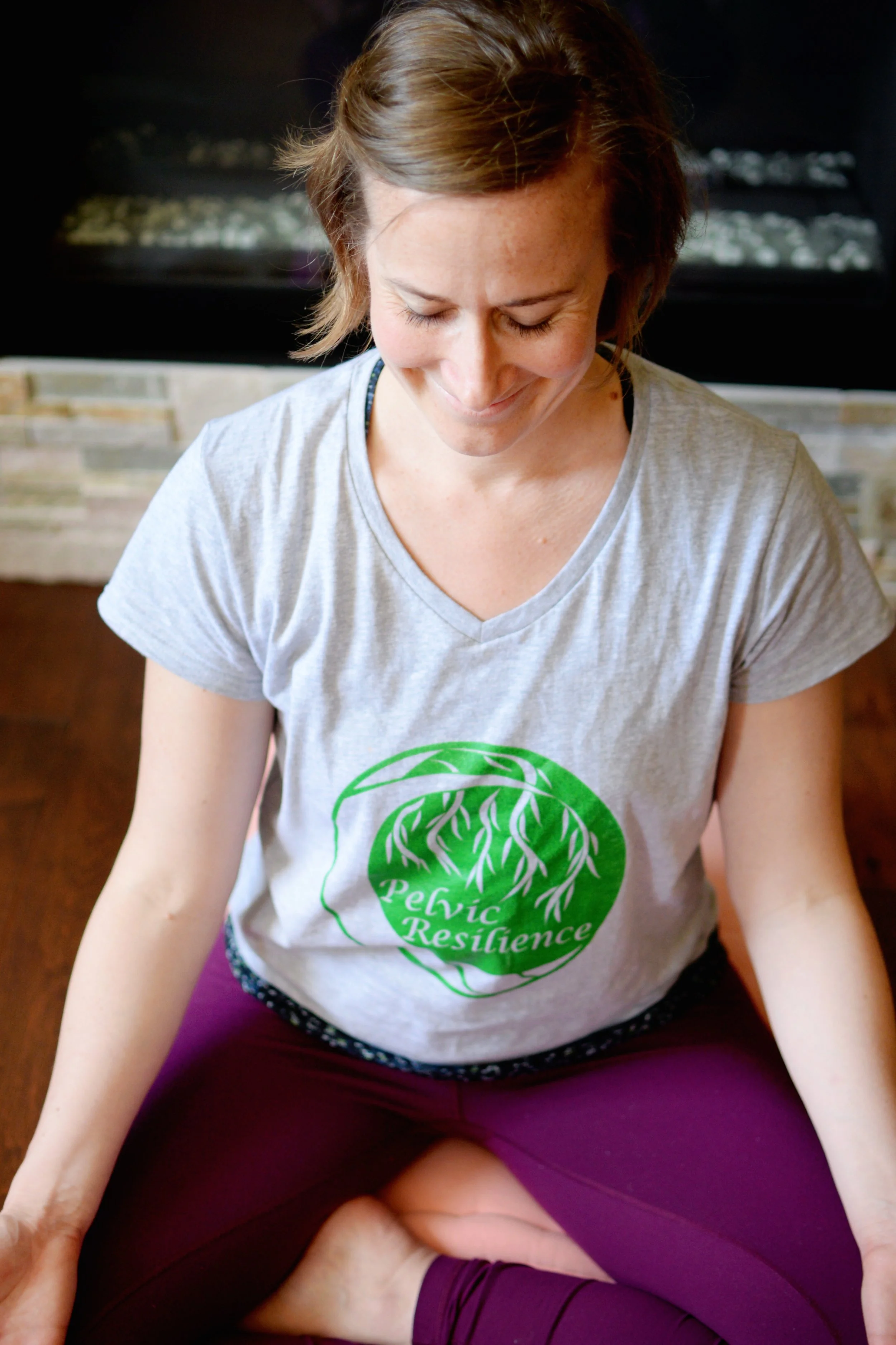 Lara Desrosiers of Pelvic Resilience practicing meditative breathing, sitting cross-legged on a yoga mat, wearing a gray t-shirt with green text and purple leggings, and smiling with her head tilted downward.
