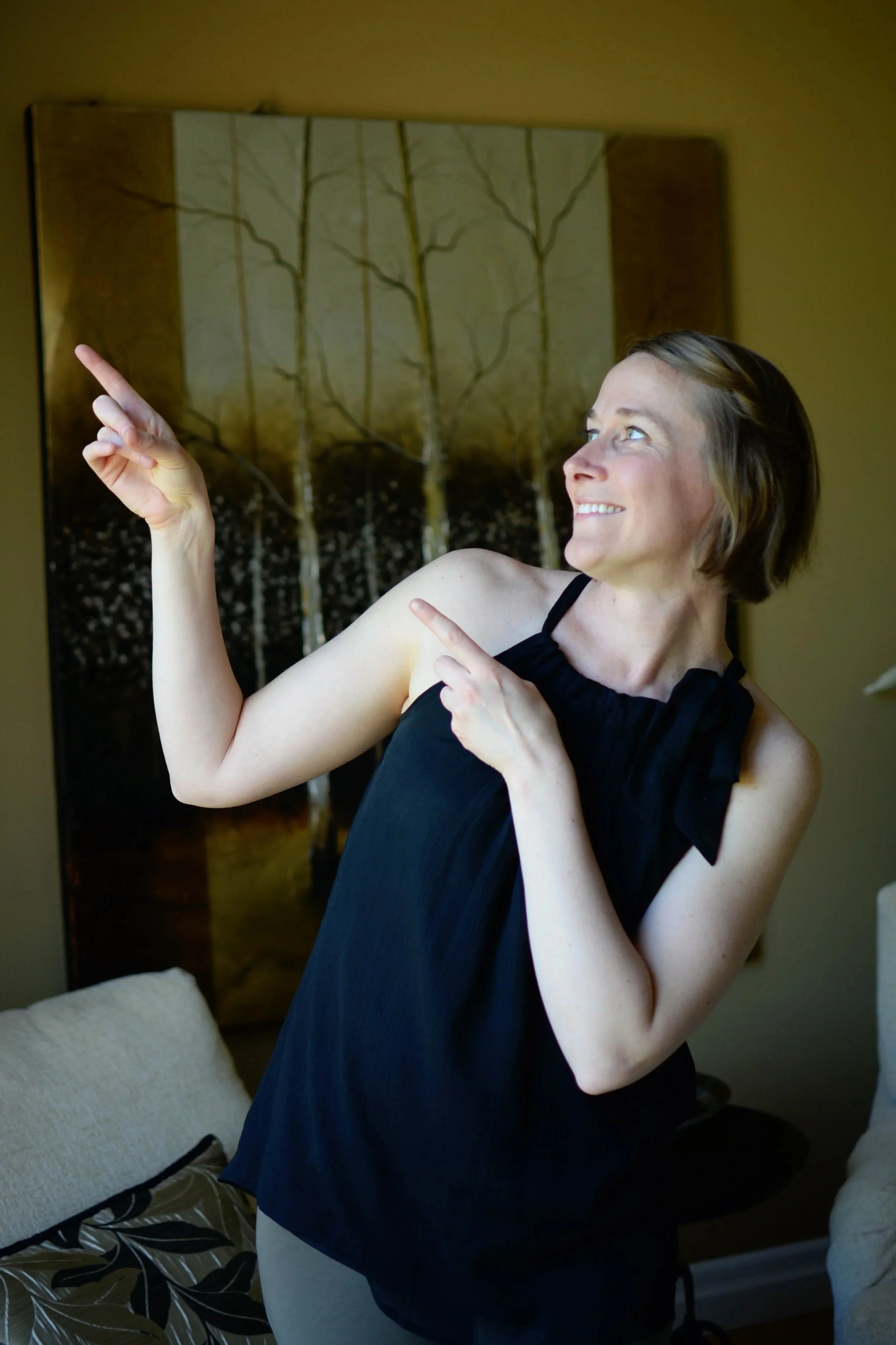 Woman with short brown hair in a black sleeveless top pointing and smiling indoors.