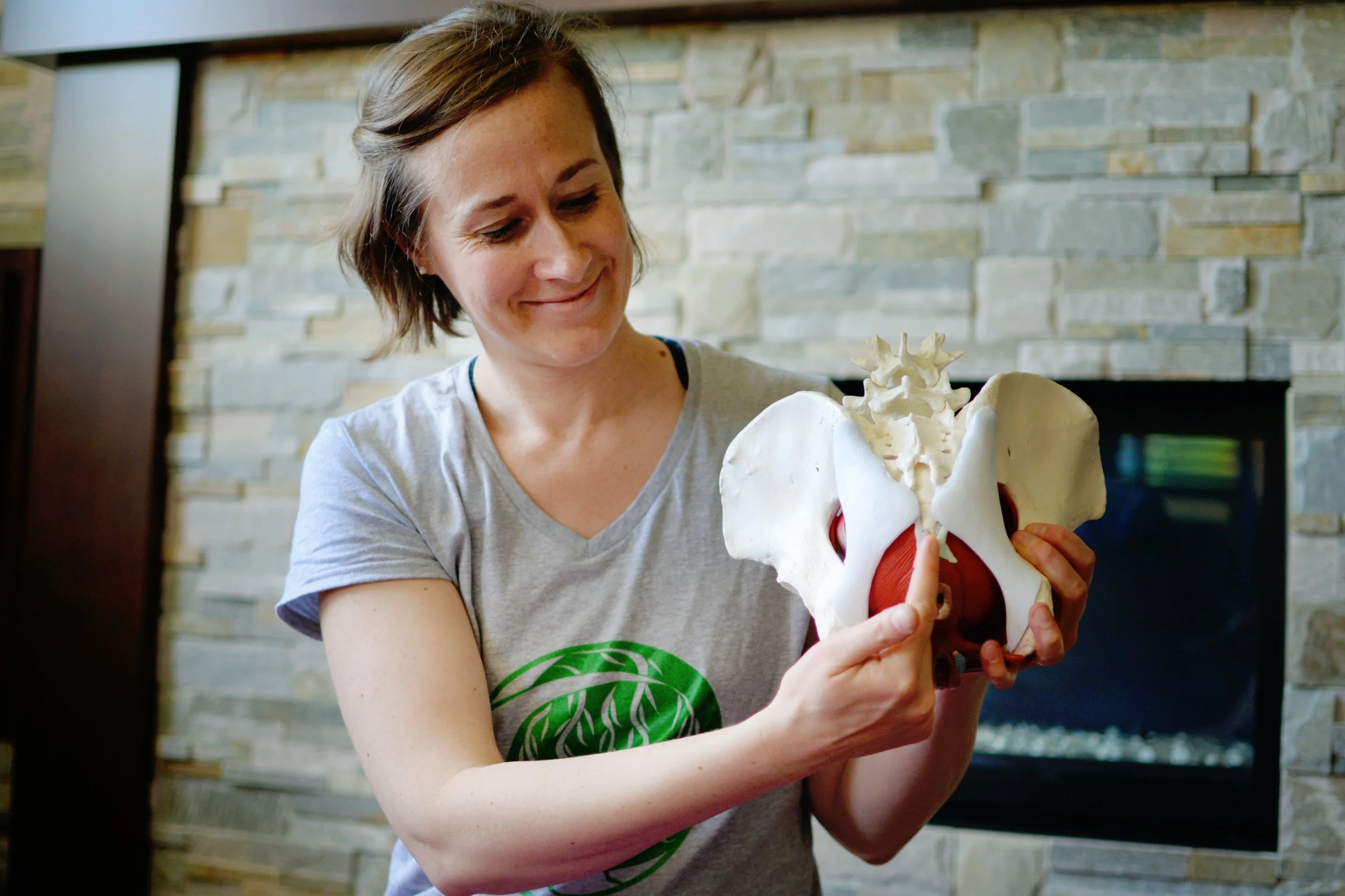 Lara Desrosiers of Pelvic Resilience holding a human pelvis anatomical model in front of a stone fireplace for pelvic health occupational therapy mentorship and education.