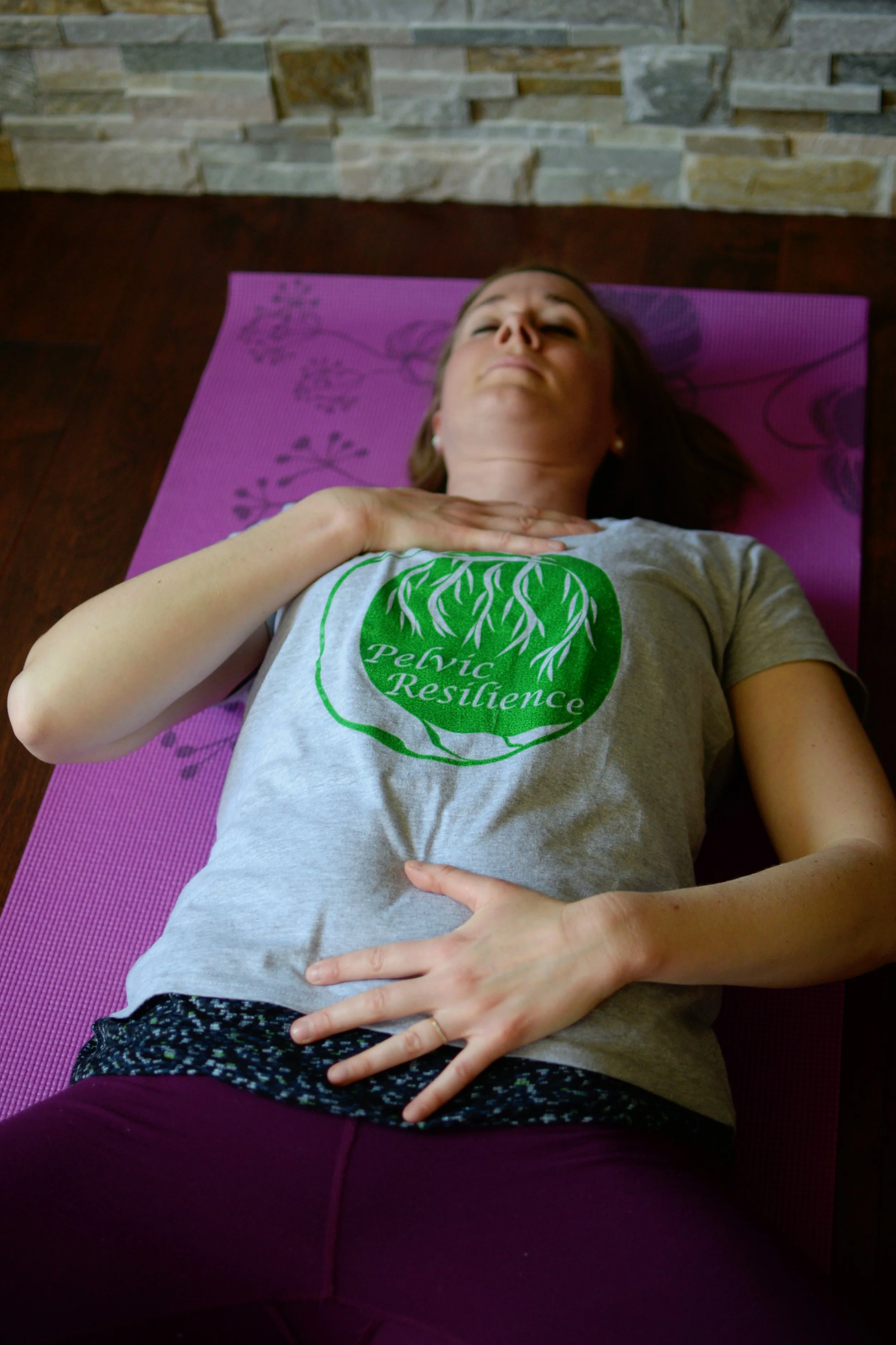 Lara Desrosiers of Pelvic Resilience lying on a purple yoga mat, practicing a breathing or relaxation exercise, wearing a grey t-shirt with a green logo that reads 'Pelvic Resilience' and a tree graphic, with her hand on her chest and stomach.