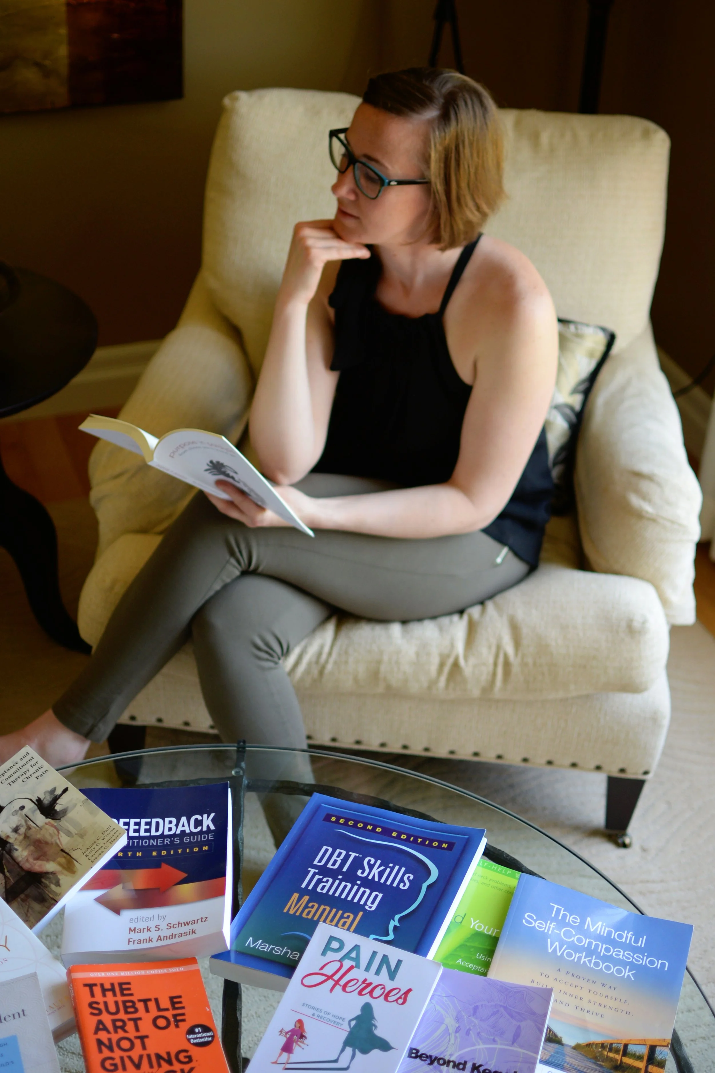 Lara Desrosiers of Pelvic Resilience with short hair and glasses sitting on a beige armchair, holding a book and looking pensively in relation to occupational therapy mentorship and continuing education for pelvic health and mental health.
