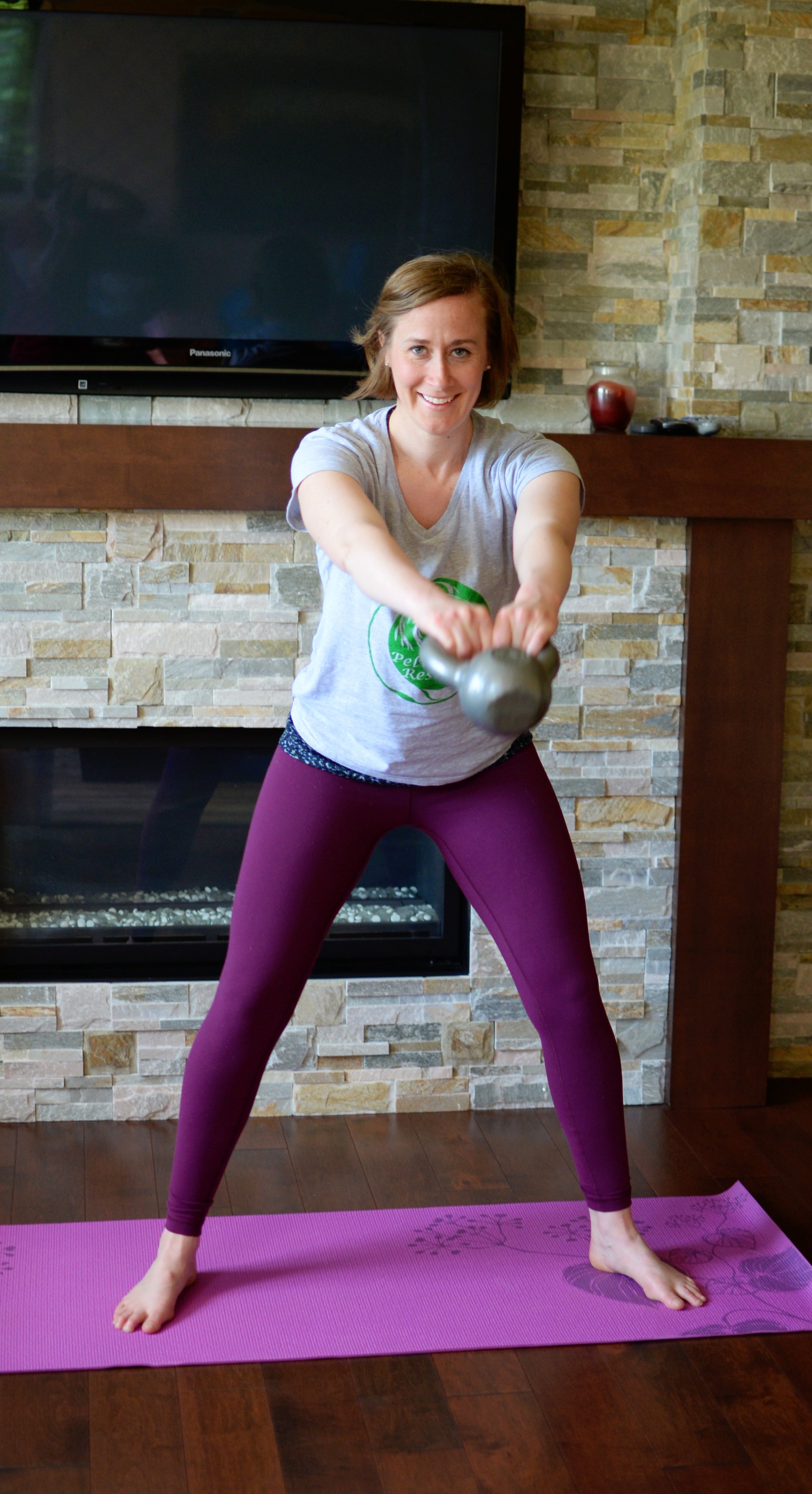 Lara Desrosiers of Pelvic Resilience doing a kettlebell exercise indoors on purple yoga mat in front of a stone fireplace with a television above.