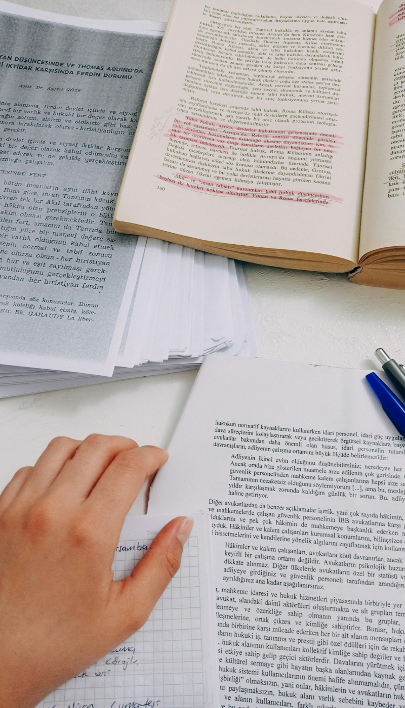 Open book with highlighted text, stack of papers, and a person's hand on a desk with pens and a notepad representing the Canadian Occupational Therapy Association's position statement on pelvic health occupational therapy.